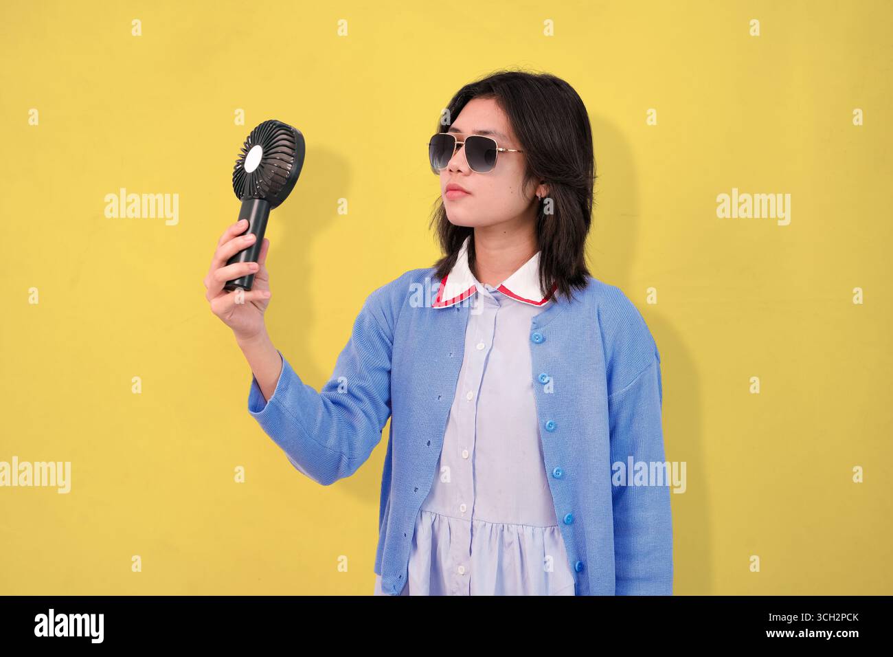 Una giovane donna sicura di sé che si raffredda con un ventilatore portatile mentre indossa gli occhiali da sole Foto Stock