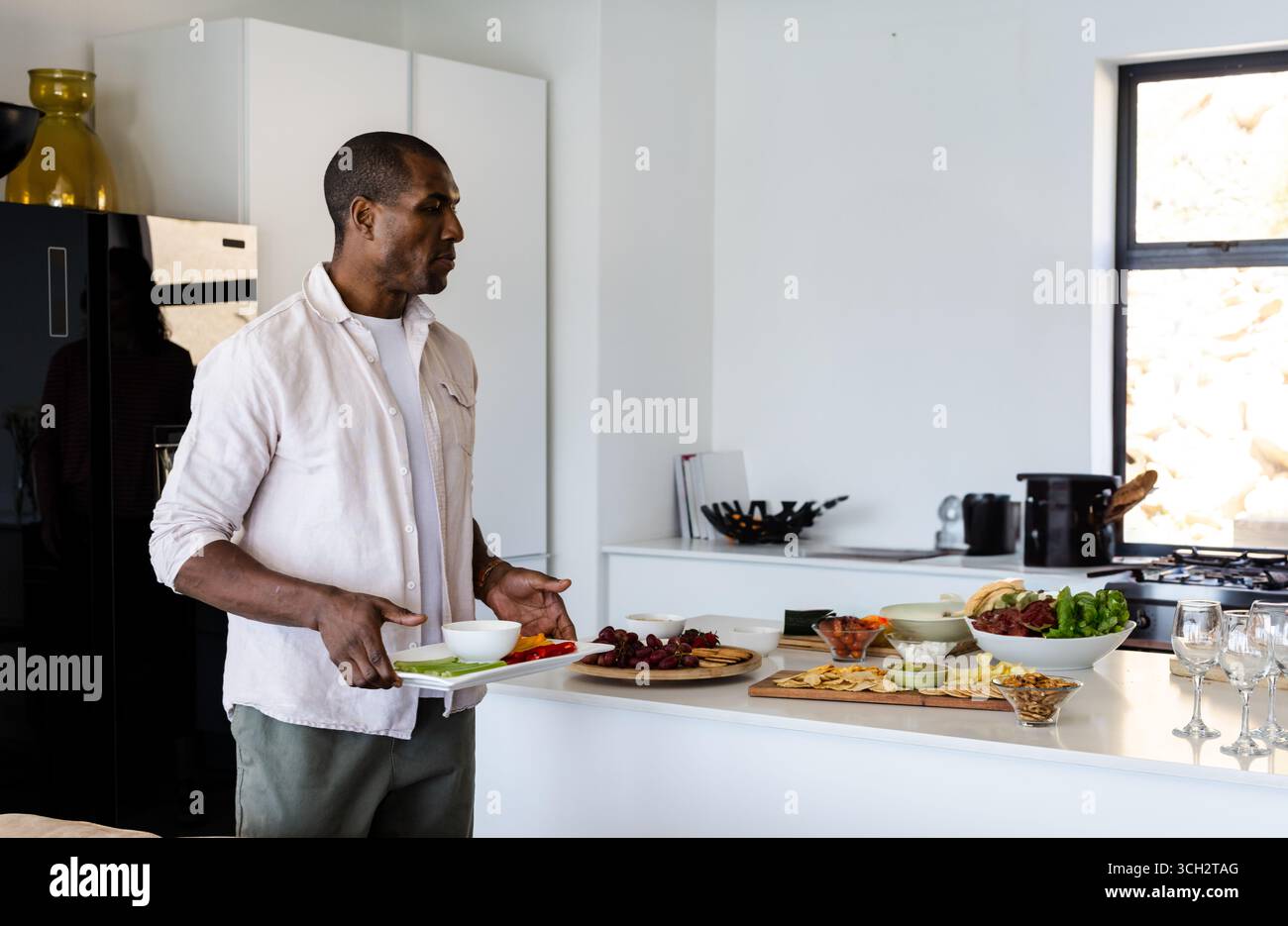 Uomo afro-americano con vassoio bianco in cucina moderna e ciotole assortite sull'isola Foto Stock