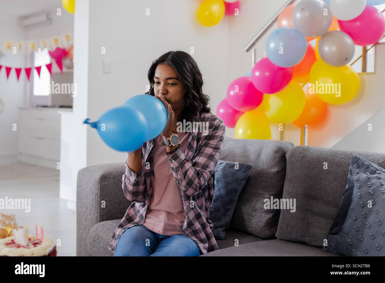 Donna che gonfia il palloncino blu sul divano grigio nel soggiorno decorato, con torta di compleanno e grappolo Foto Stock
