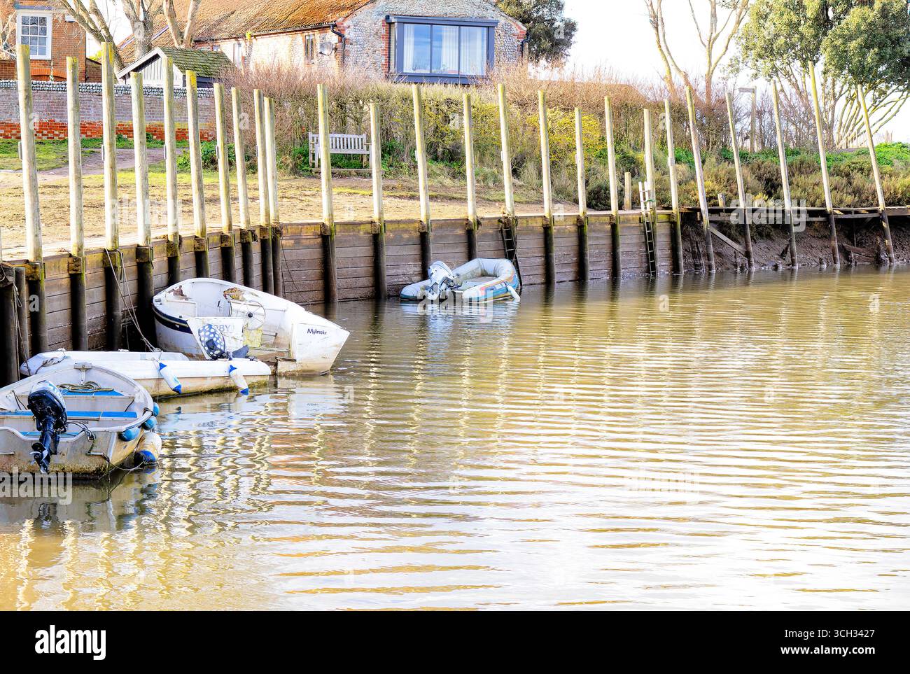 Una piccola barca riposa tranquillamente lungo un molo in legno lungo il fiume, che si riflette nelle acque calme sotto il verde delle sorgenti. Foto Stock