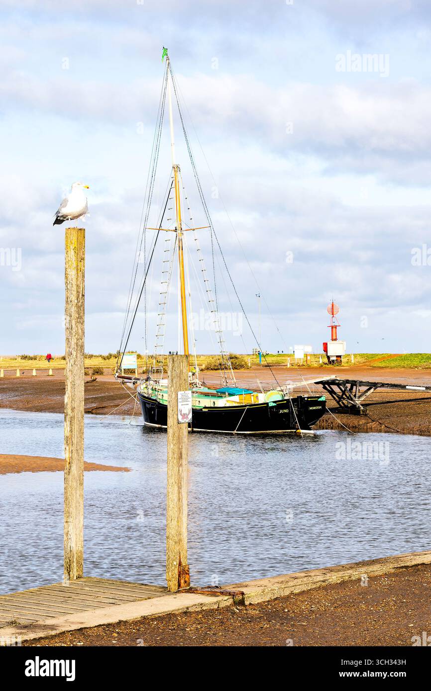 Una tradizionale barca a vela chiamata Juno riposa tranquillamente con la bassa marea nel tranquillo porto di Morston, sotto un cielo luminoso disseminato di morbide nuvole. Foto Stock