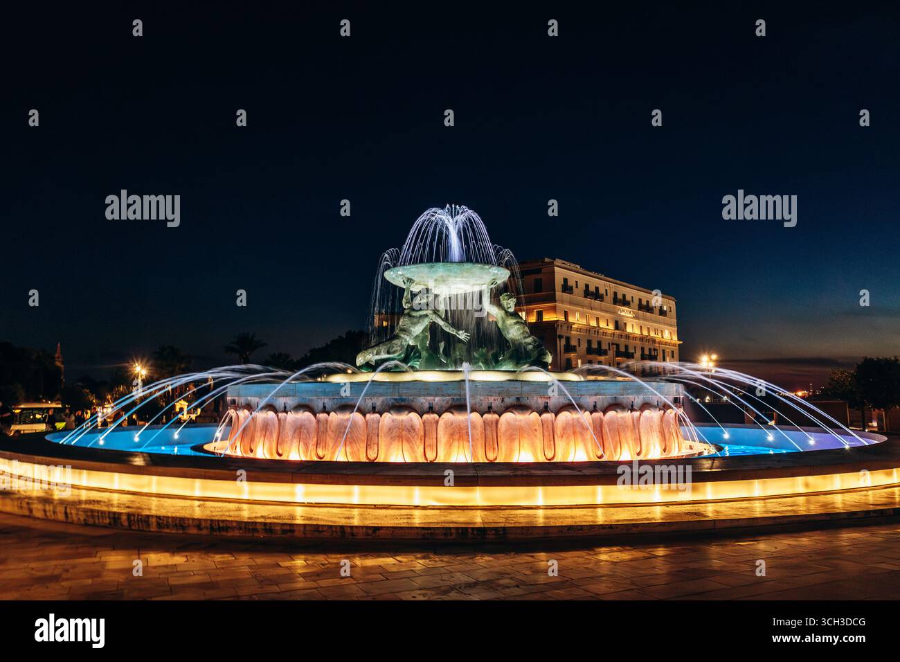 La Fontana del Tritone illuminata di notte, con tre figure di bronzo che tengono in mano un grande bacino all'ingresso della città di la Valletta Foto Stock