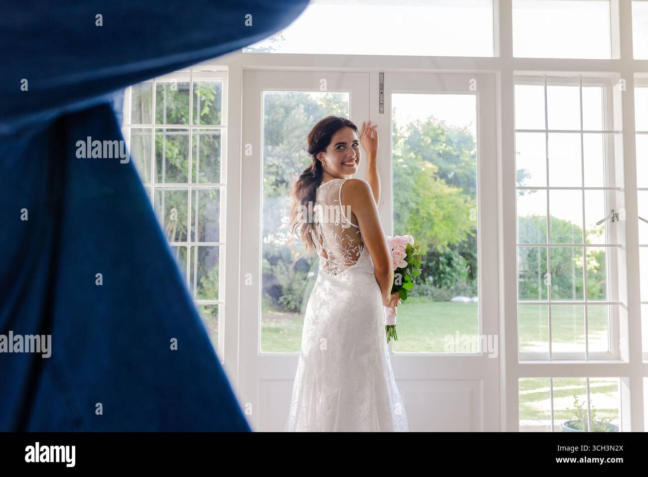 Sposa in elegante abito in pizzo sorridente tenendo il bouquet vicino alla finestra illuminata dal sole Foto Stock
