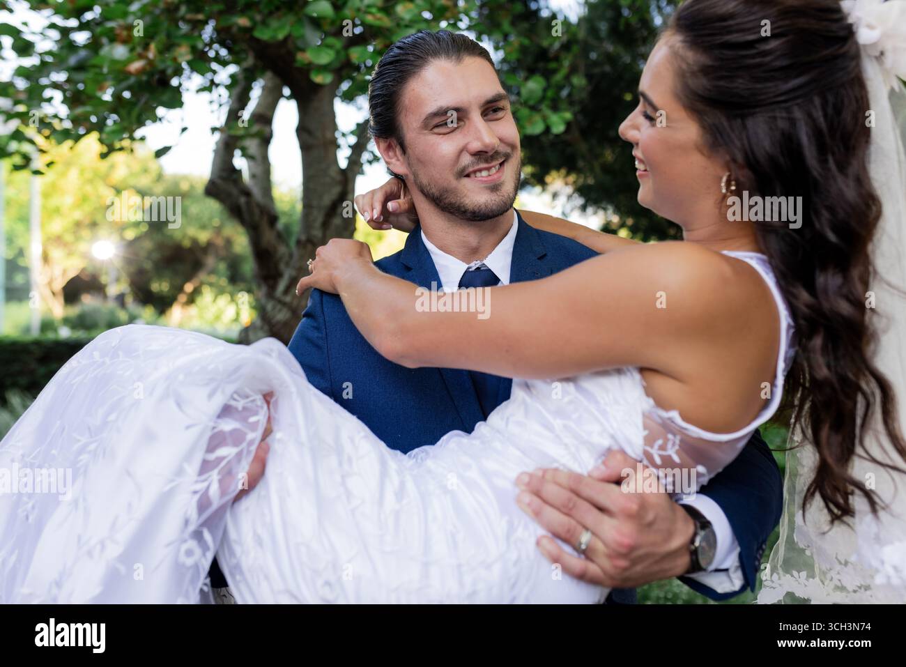 Sposo che porta con gioia la sposa in giardino, sorridendo e guardando l'un l'altro Foto Stock
