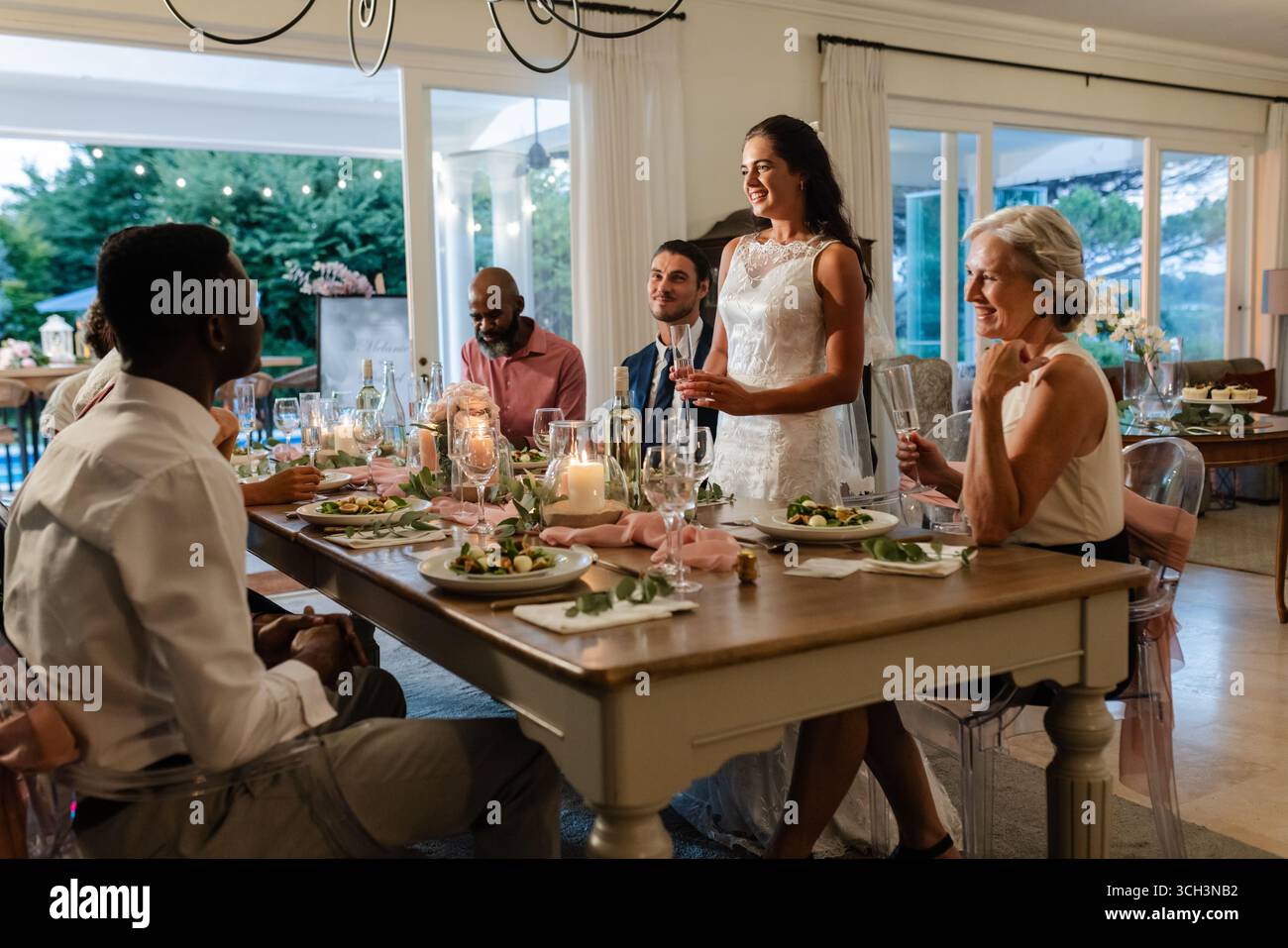 Sposa in abito bianco che parla con gioia agli ospiti a un elegante tavolo da pranzo all'interno Foto Stock