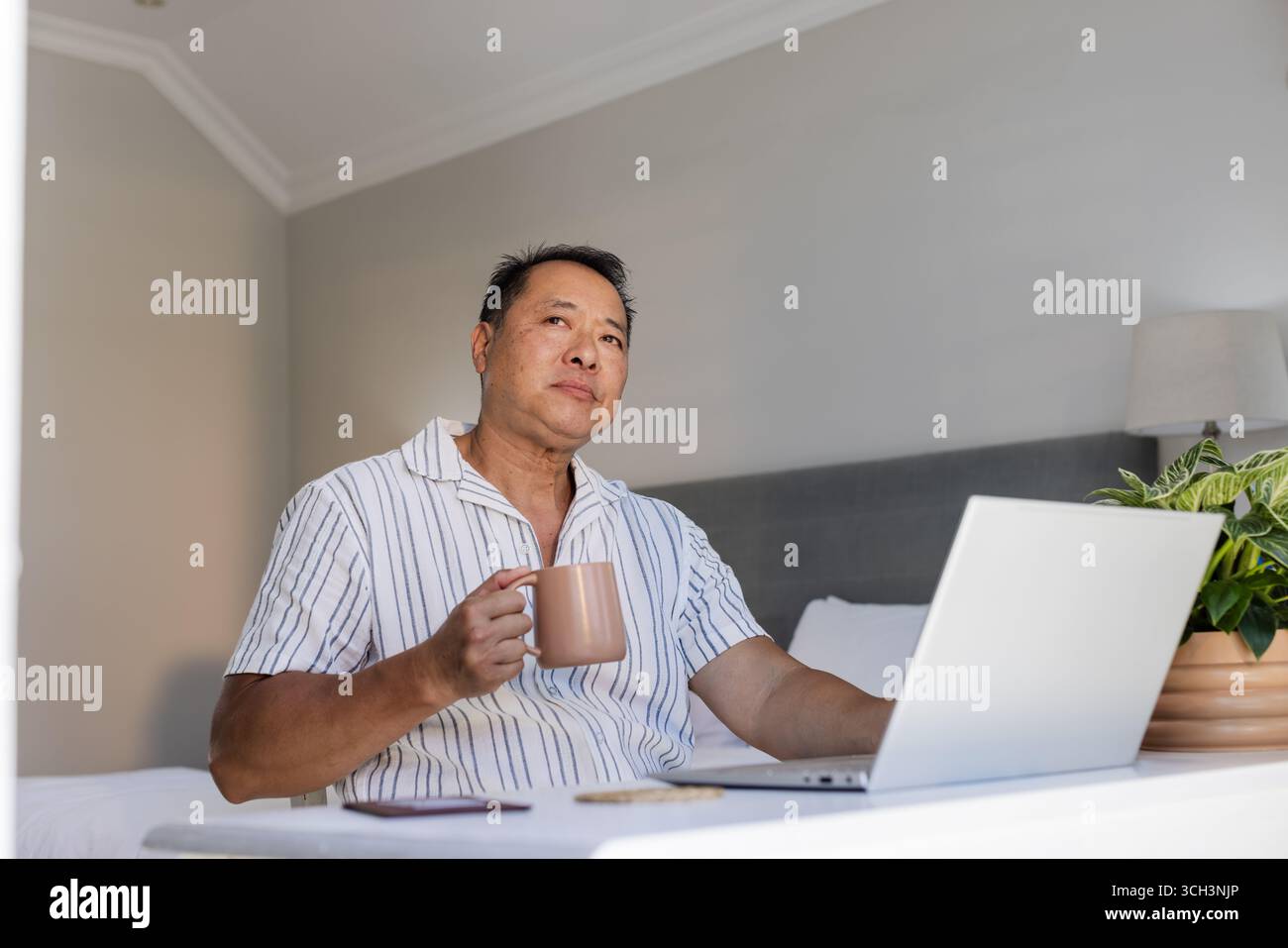 Un uomo asiatico anziano che si gusta un caffè mentre usa un notebook a casa, con un aspetto premuroso Foto Stock