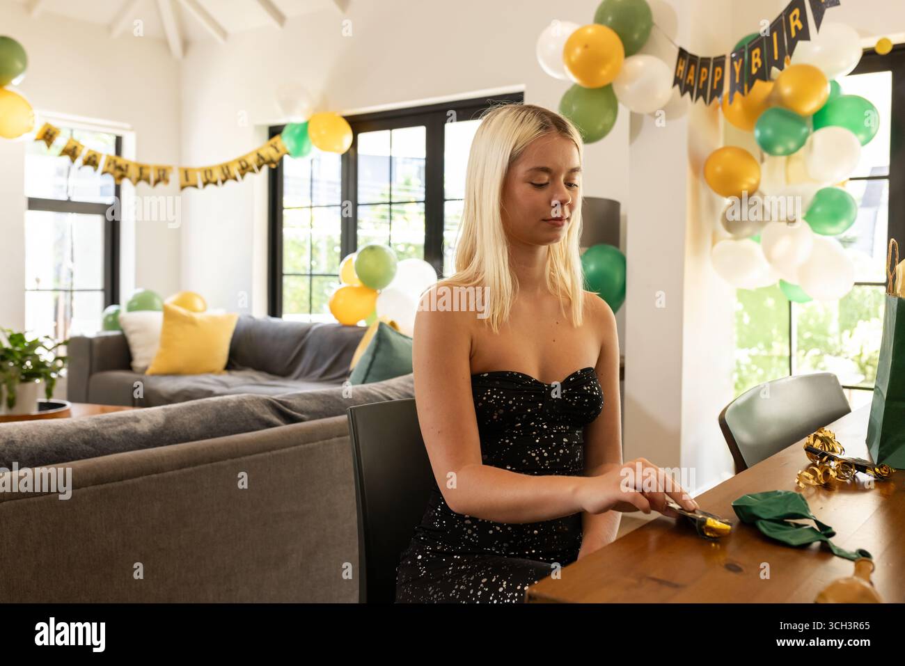 Donna seduta al tavolo che svela il regalo di compleanno nel soggiorno con palloncini, striscioni, spazio per le copie Foto Stock