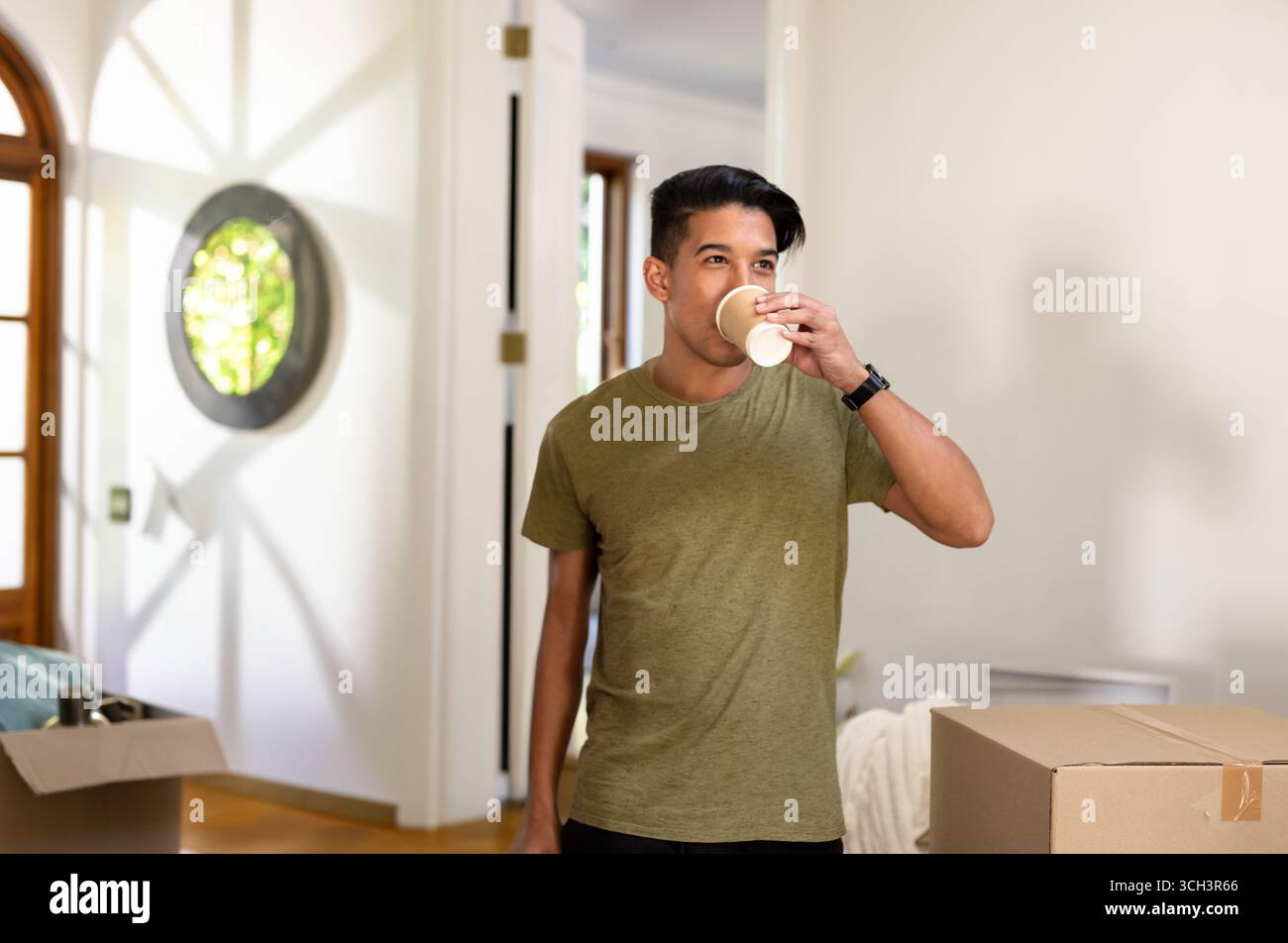 Giovane asiatico sorseggiando da una tazza di carta nell'ingresso di casa, con scatole mobili e ombre radiali Foto Stock