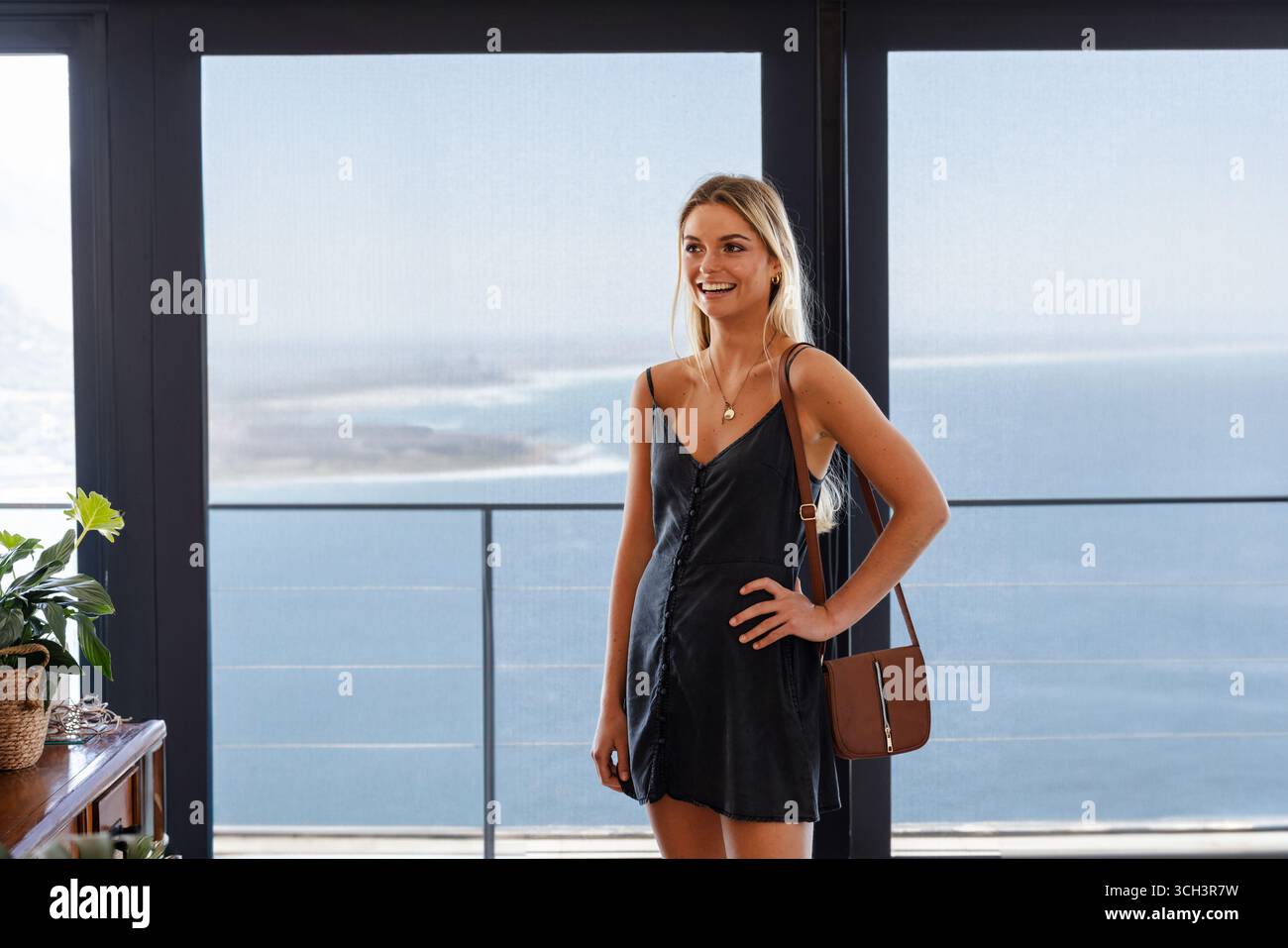 Donna che indossa un abito nero che posa davanti a una porta di vetro e guarda l'oceano accanto a una piantatrice intrecciata che porta una borsa Foto Stock