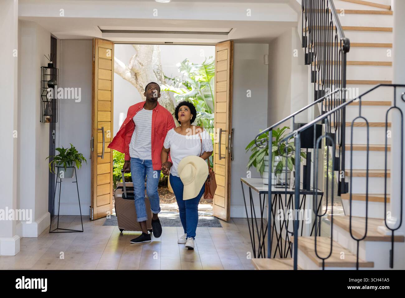 Coppia afroamericana che entra in casa moderna portando valigia e cappello, sembrando rilassata e felice Foto Stock