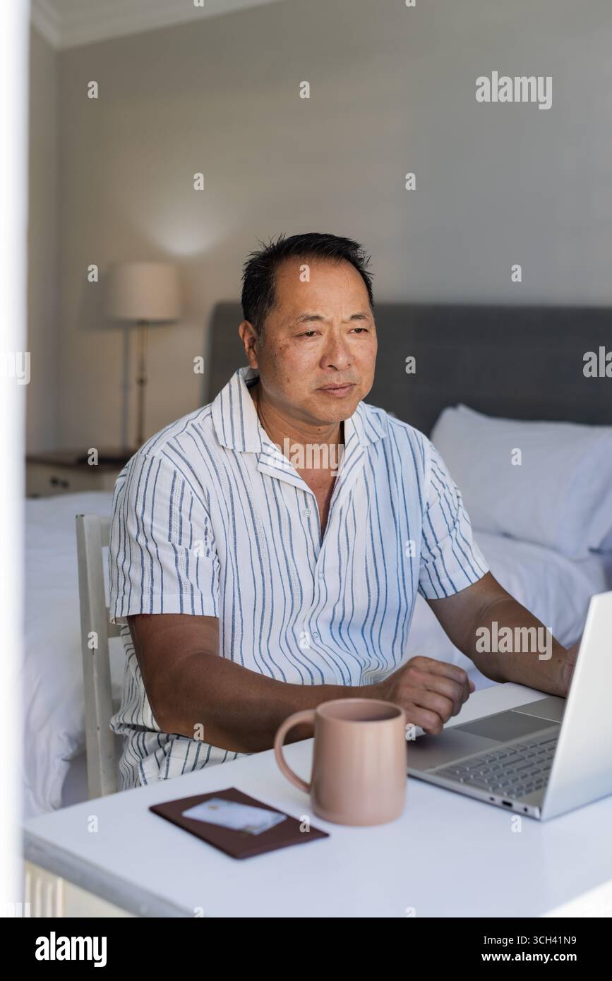 Uomo asiatico anziano che utilizza un computer portatile a casa, concentrandosi sul lavoro con caffè nelle vicinanze Foto Stock