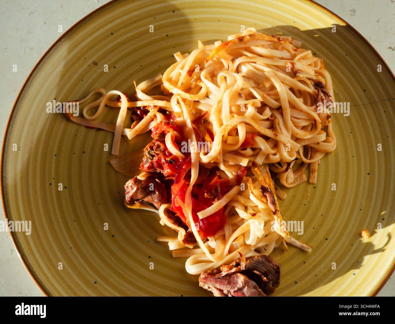 La pasta fatta in casa, condita generosamente con carne salata e ricca salsa di pomodoro, che riposa su un piatto giallo vibrante, offre un pasto delizioso e soddisfacente Foto Stock
