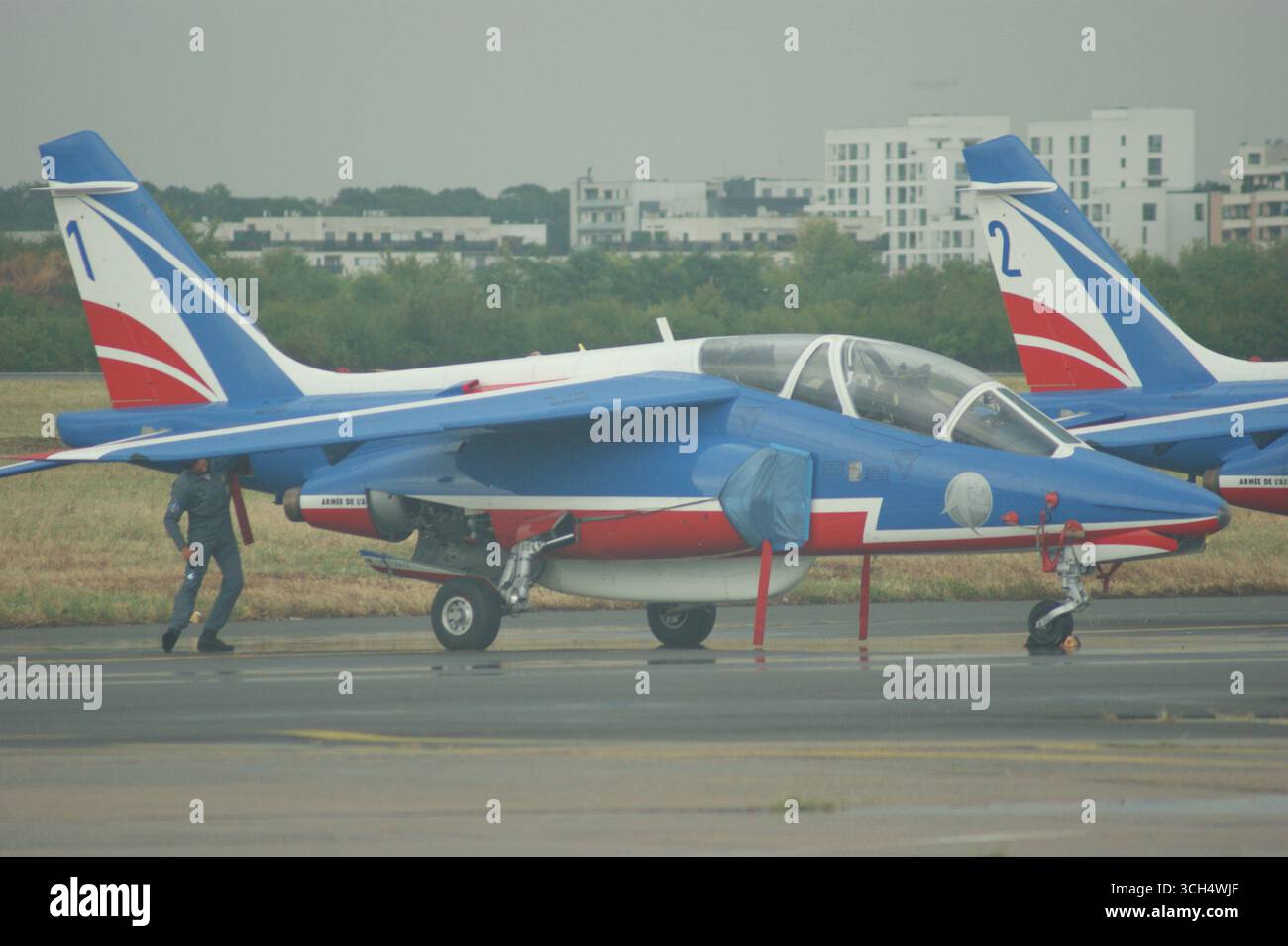 Un Alpha Jet dell'aeronautica francese in livrea blu, bianca e rossa è parcheggiato su un asfalto, con una persona in tuta di volo che cammina nelle vicinanze e negli edifici urbani. Foto Stock