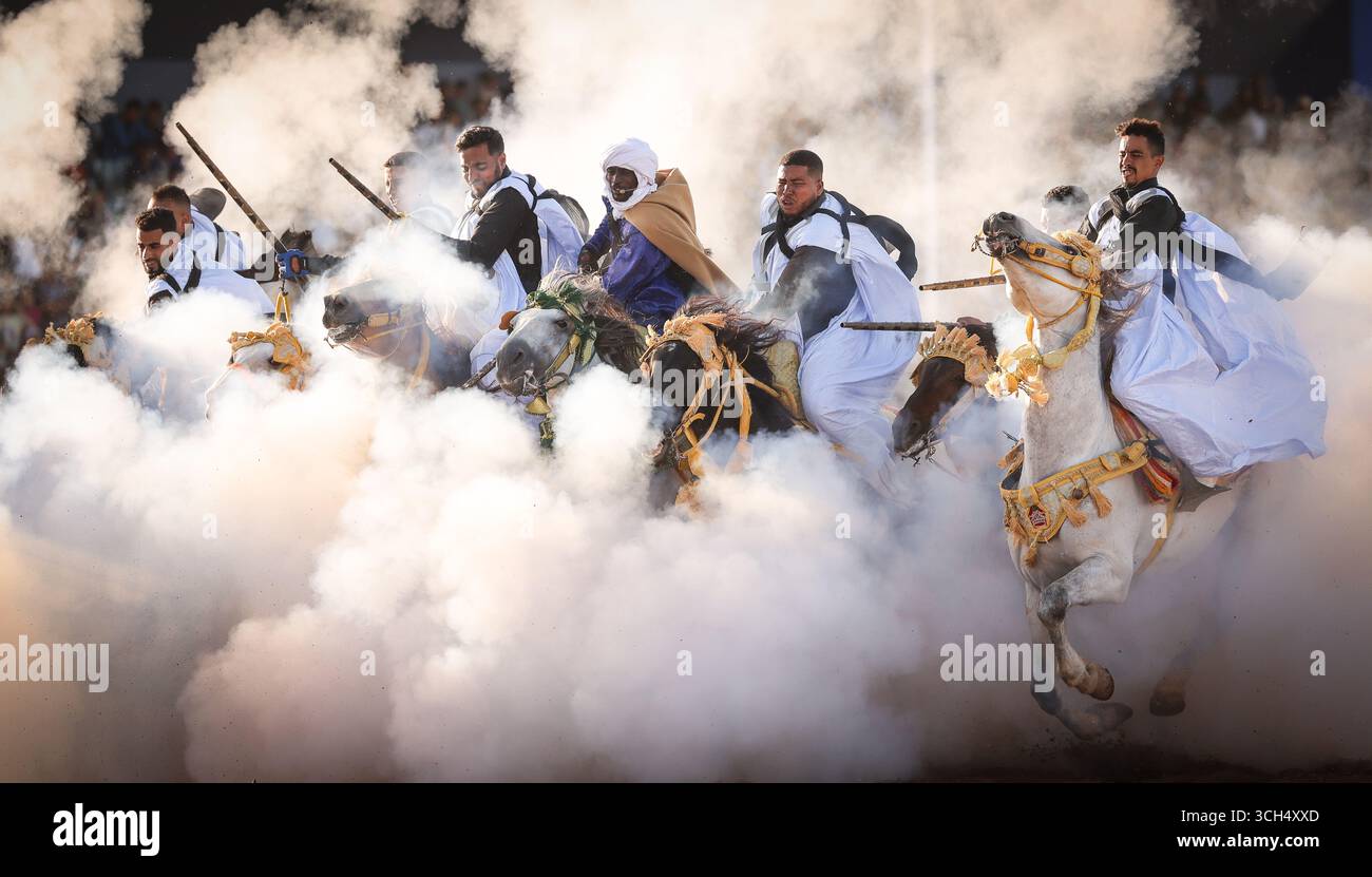 Una storia di coraggio, unità e eredità catturata in un unico fotogramma.Riders in colorati costumi marocchini al galoppo su cavalli arabi in uno spettacolo Fantasia Foto Stock