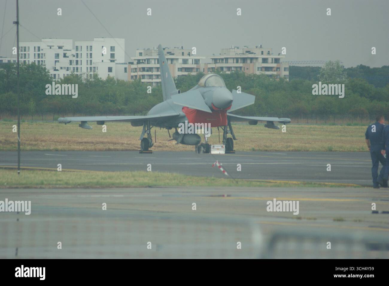 Un jet Eurofighter Typhoon è parcheggiato su un asfalto, sullo sfondo di moderni edifici urbani, che rappresentano la tecnologia militare e la vita urbana. Foto Stock