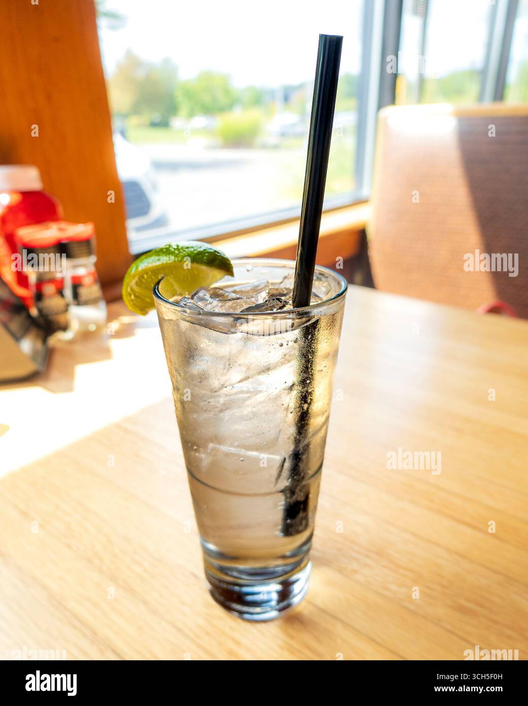 Un alto gin tonico con un cuneo di lime e una paglia scura siede su un tavolo di legno, illuminato dalla luce del sole. Foto Stock