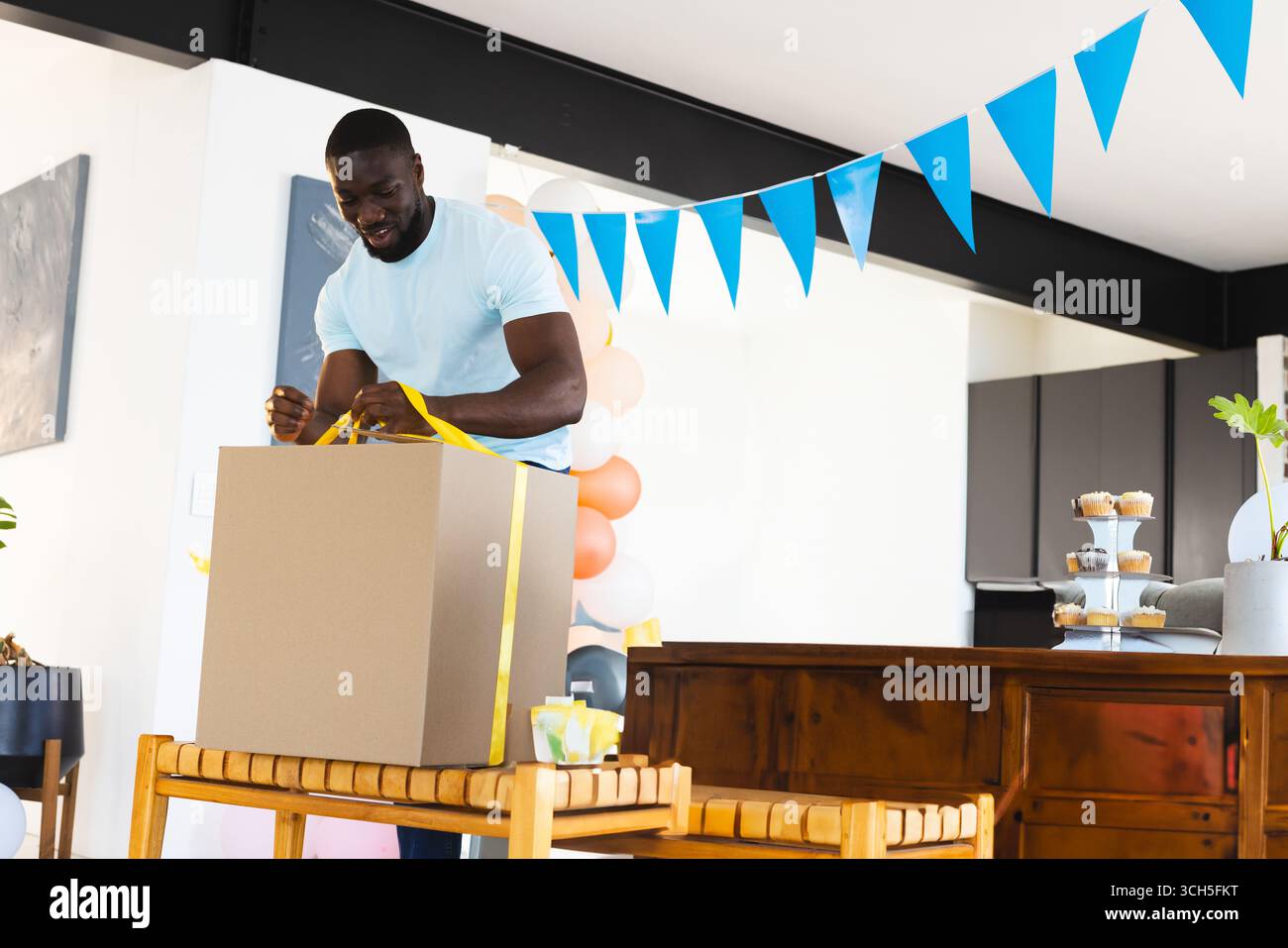 Scatola regalo per uomo afroamericano in sala da pranzo, con nastro giallo, palloncini e cupcake Foto Stock