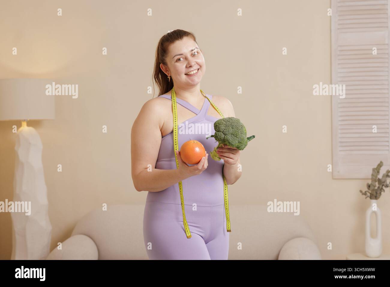 Donna sorridente in abbigliamento sportivo che tiene broccoli e pompelmo, mostrando sane abitudini alimentari, dieta e motivazione per la perdita di peso Foto Stock