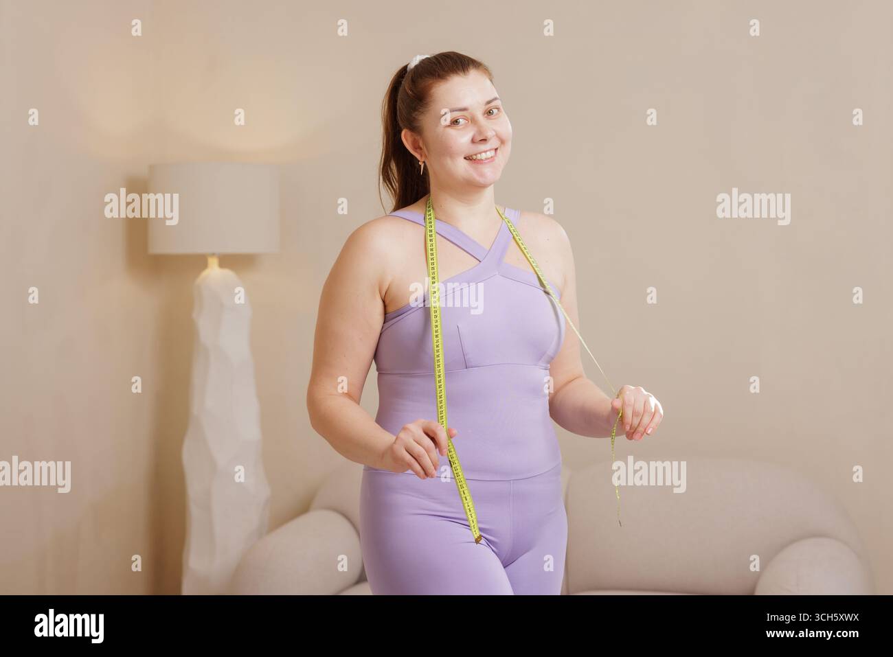 Donna sorridente più taglia in abbigliamento sportivo che tiene il metro a nastro, mostrando il progresso di perdita di peso, abitudini sane e positività del corpo Foto Stock