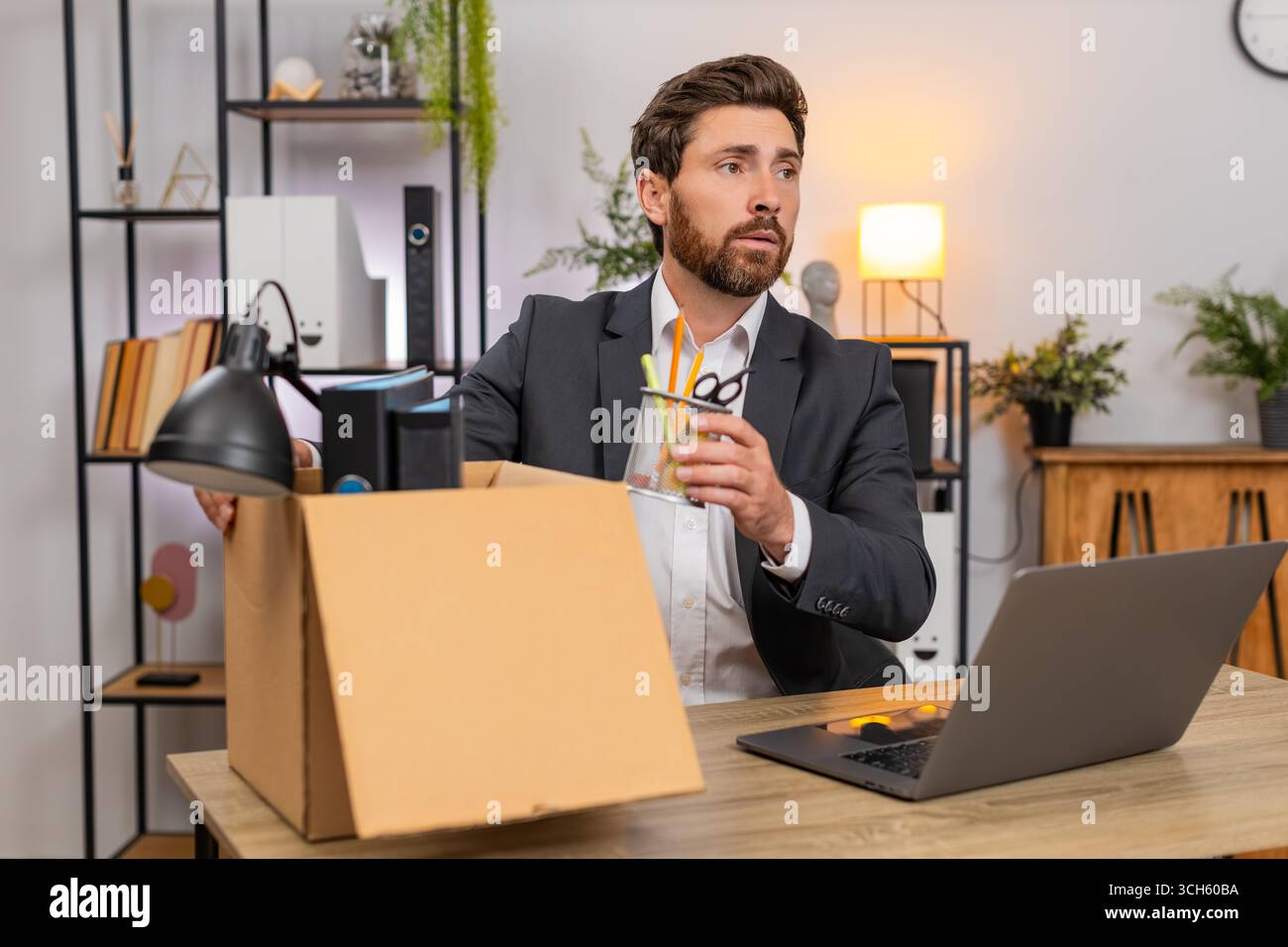 Un uomo d'affari di mezza età in ufficio porta con cura gli oggetti nella scatola e sbircia con nostalgia. Il freelance alla scatola delle etichette del tavolo chiude il notebook e sorride pronto per il prossimo passo in avanti. Foto Stock