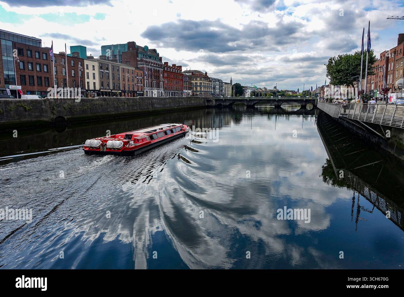 Il tour in barca rosso attraversa il fiume Liffey a Dublino, Irlanda, con riflessi di nuvole, edifici storici lungo la banchina e Grattan Bridge in lontananza Foto Stock