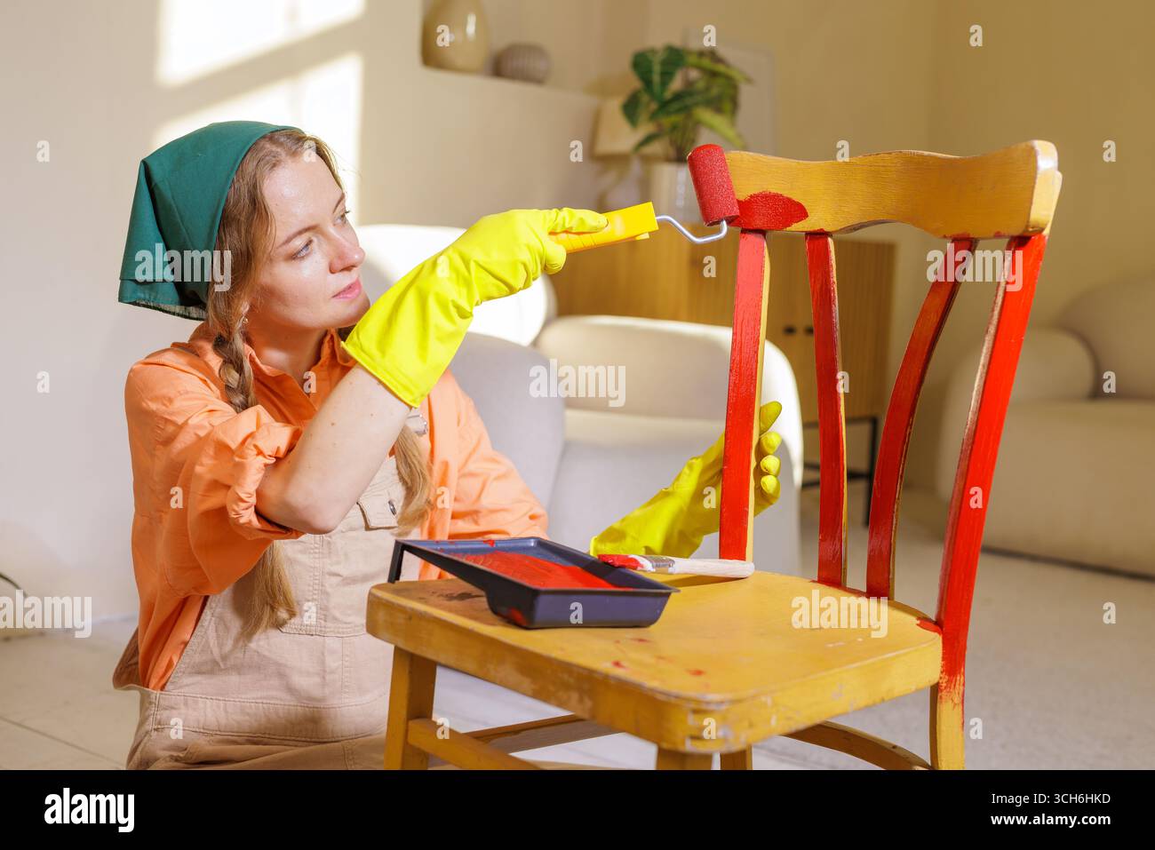Donna in guanti che dipinge una sedia in legno con rullo di vernice rosso a casa, concetto di ristrutturazione fai da te e upcycling di mobili Foto Stock
