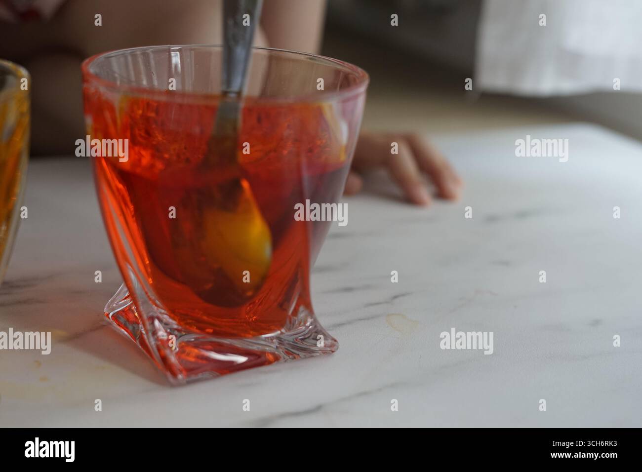 Una vivace bevanda arancione-rossa mescolata con un cucchiaio in un bicchiere elegante, che evoca un gusto rinfrescante e un'atmosfera calda per incontri o momenti informali Foto Stock Una vivace bevanda arancione-rossa mescolata con un cucchiaio in un bicchiere elegante, che evoca un gusto rinfrescante e un'atmosfera calda per incontri o momenti informali Foto Stock