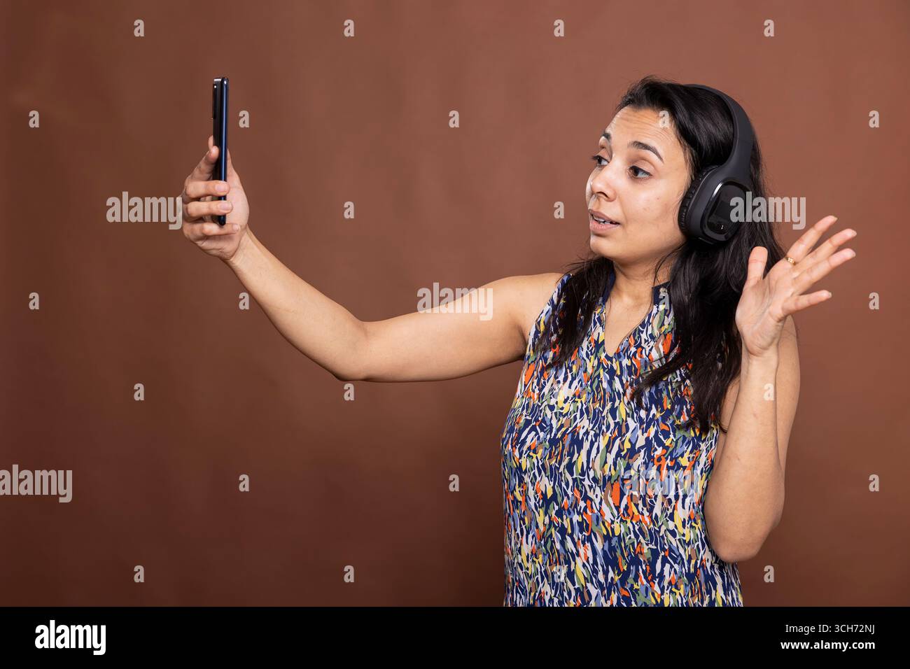 Bella modella femminile che indossa le cuffie si aggancia al cellulare mentre sorride. Graziosa donna indiana con cuffie wireless, gode di una videochiamata con la famiglia, in piedi su uno sfondo isolato. Foto Stock