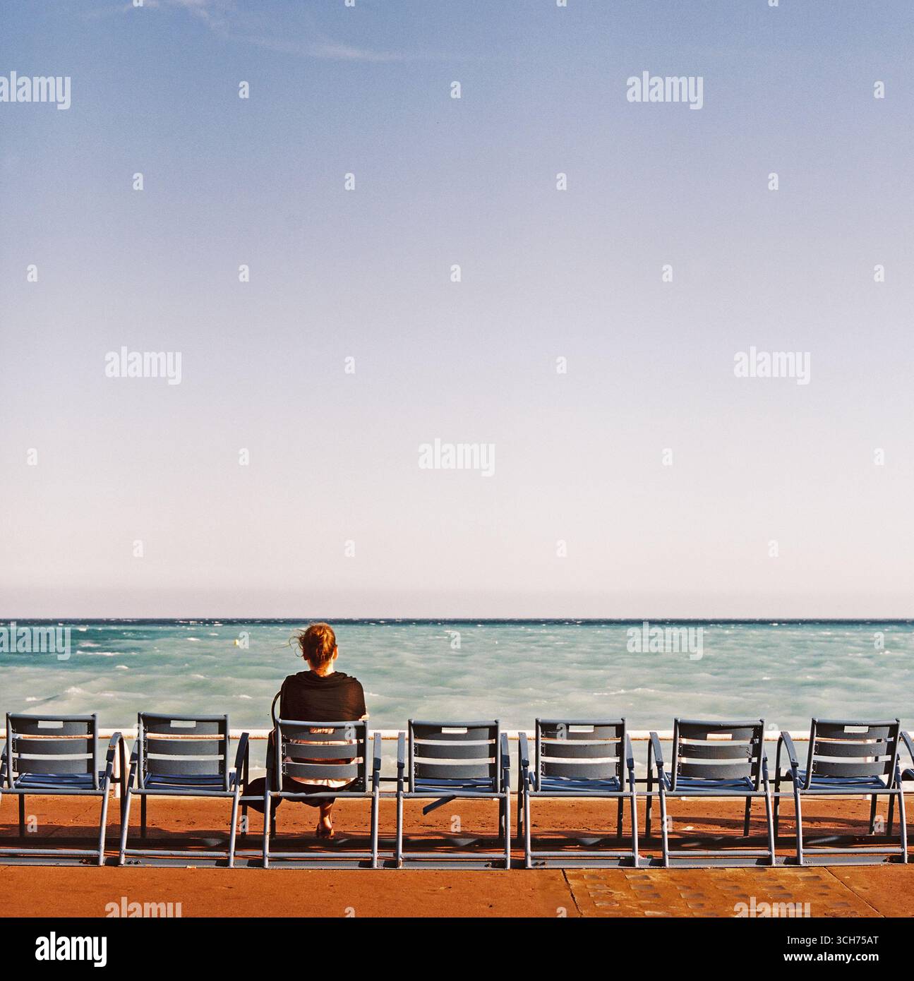 Donna seduta sulla spiaggia e guardando il mare, vista da dietro. Un momento tranquillo di solitudine e riflessione, fotografato su film analogici in Francia. Foto Stock