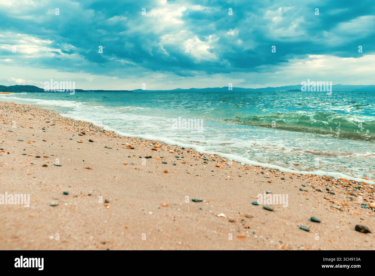 Spettacolare paesaggio marino di Policrono, Halkidiki, Grecia, che mostra onde turchesi che si lavano su una spiaggia di sabbia di ciottoli sotto un cielo lunare. Montagne lontane Foto Stock