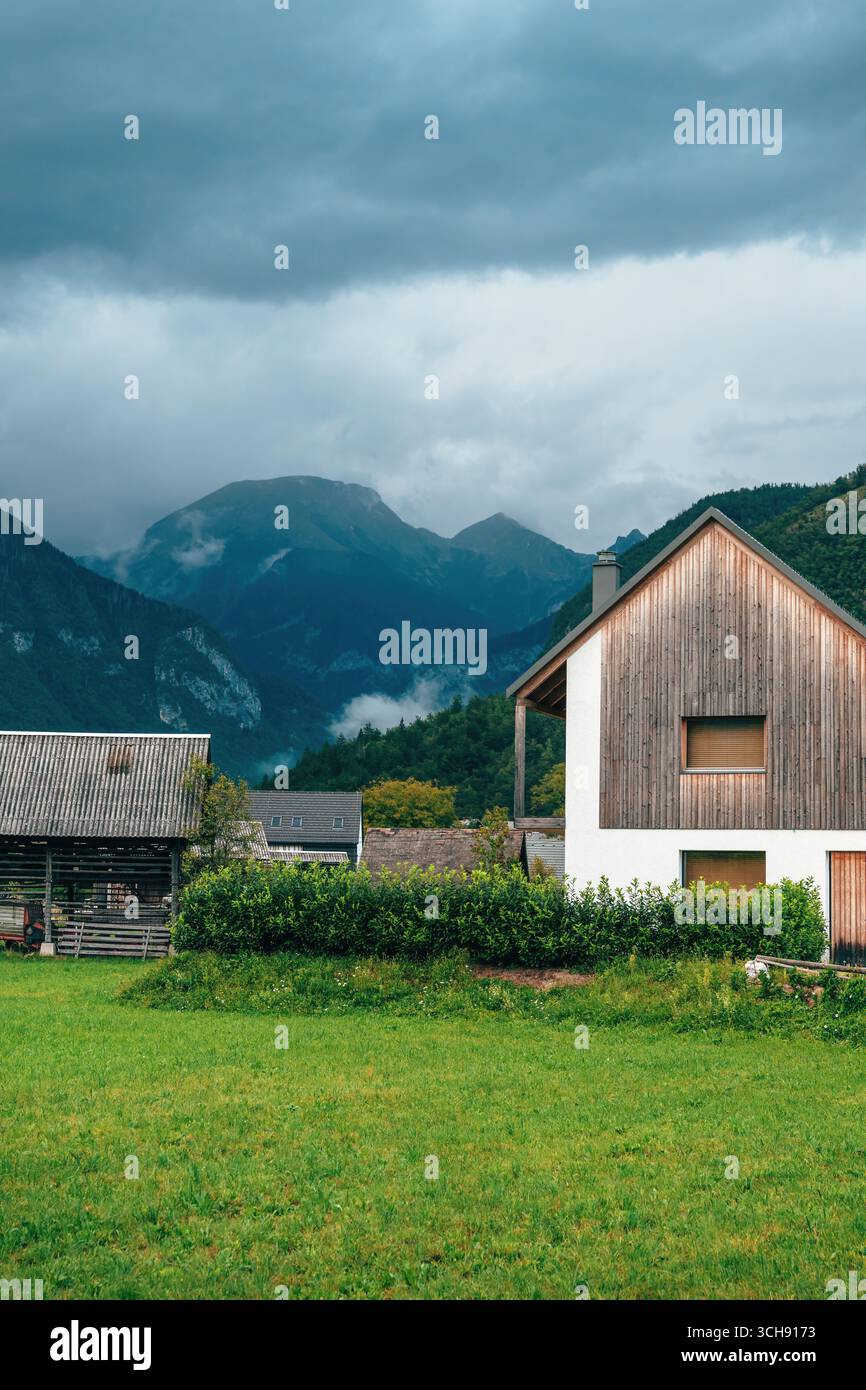Casa contemporanea sulle Alpi con facciata in legno, annidata in una valle tranquilla con montagne boscose e nuvole nebbiose. Messa a fuoco selettiva. Foto Stock