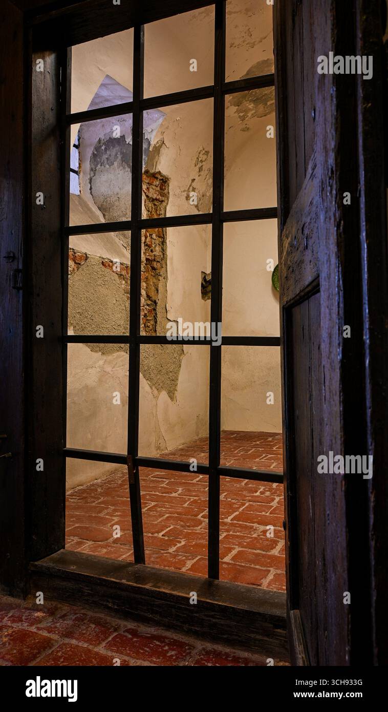 Vista di una vecchia cella della prigione con pareti in gesso sbriciolate e pavimento in piastrelle di terracotta visto attraverso barre di metallo. Foto Stock