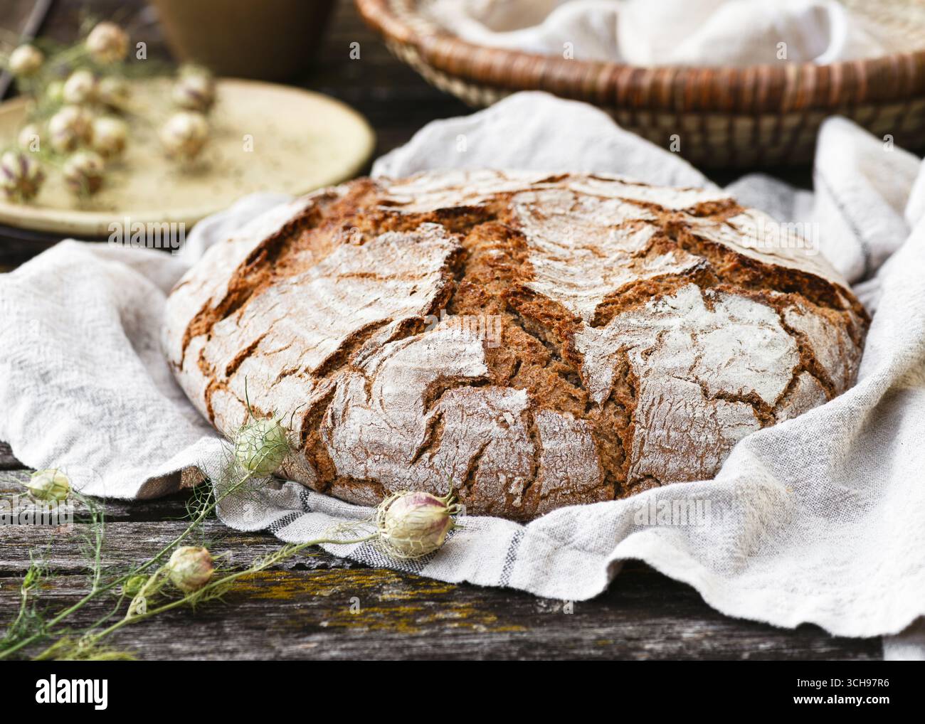 Il pane dorato e croccante si trova su un panno morbido, la sua superficie ruvida e piena di carattere. Dettagli rustici con legno, vimini e fiori Foto Stock