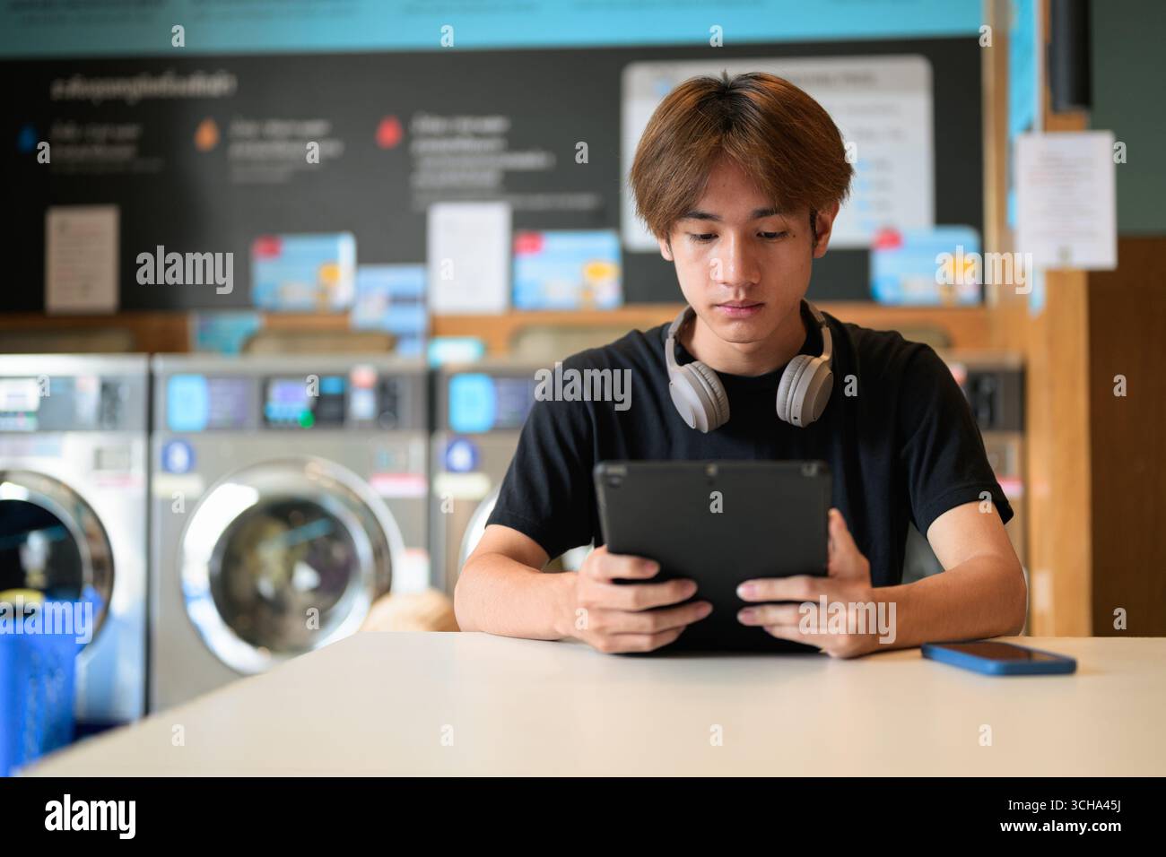 Giovane uomo thailandese seduto in una moderna lavanderia con lavatrice e con una t-shirt nera. Ritratto sincero che mostra lo stile di vita della generazione Z, le faccende quotidiane, Foto Stock