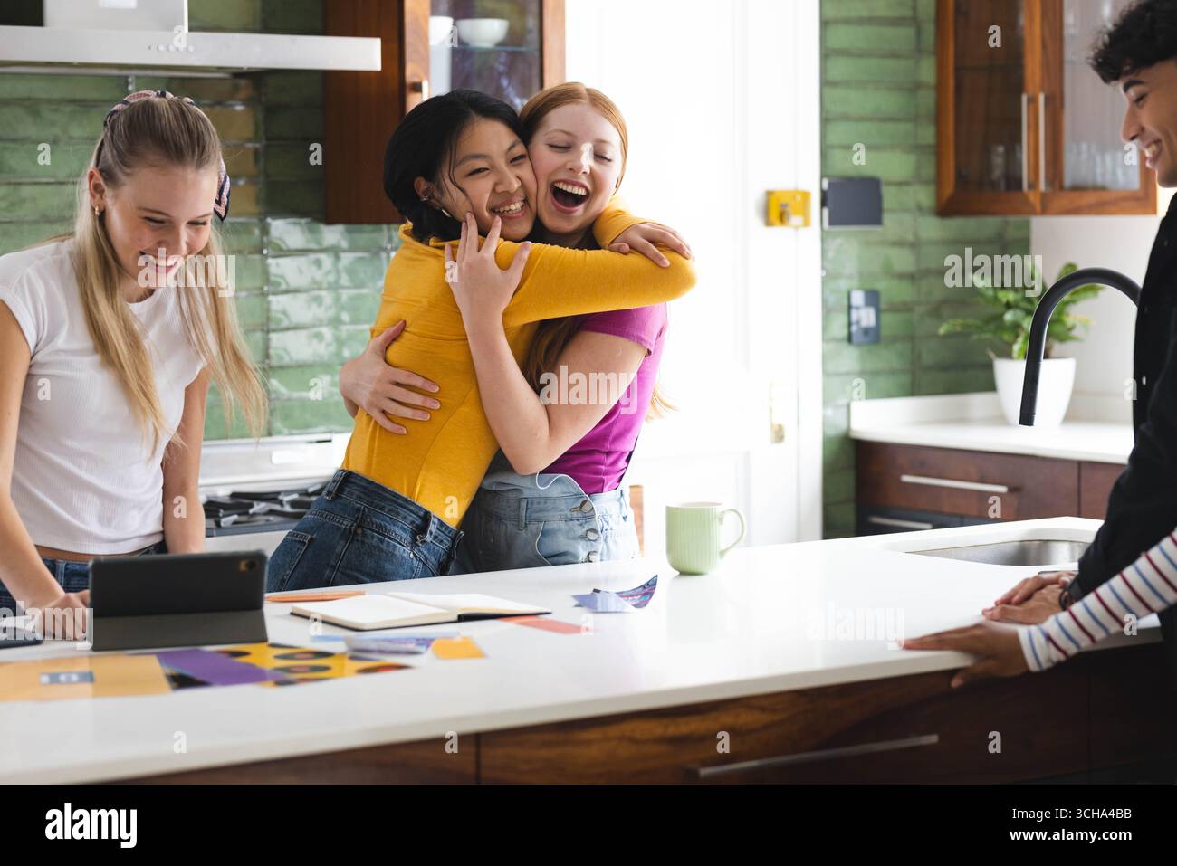 Amici diversi che festeggiano in cucina, si abbracciano e sorridono intorno a tablet e giornali, a casa. Amicizia, celebrazione, tecnologia, giovinezza, togeth Foto Stock