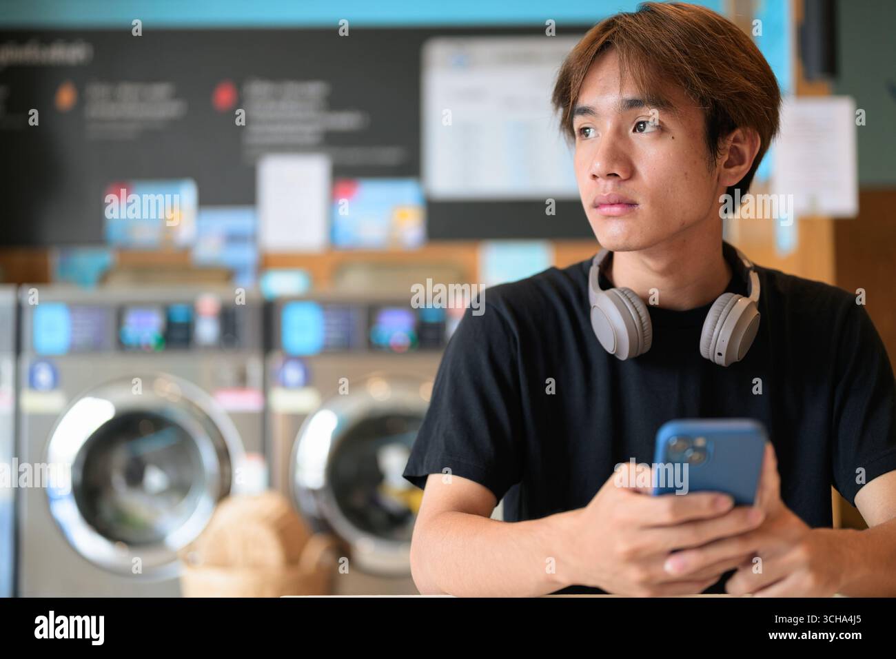 Giovane e affascinante uomo thailandese seduto in una moderna lavanderia con lavatrice che usa il telefono e indossa una t-shirt nera. Ritratto sincero che mostra la vita della Gen Z Foto Stock