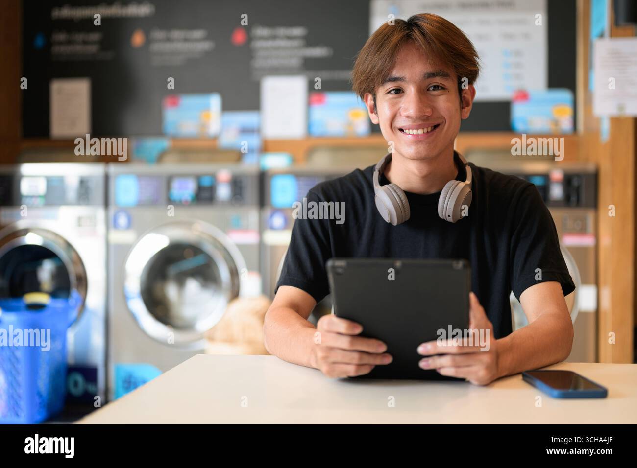 Giovane uomo thailandese seduto in una moderna lavanderia con lavatrice e con una t-shirt nera. Ritratto sincero che mostra lo stile di vita della generazione Z, le faccende quotidiane, Foto Stock