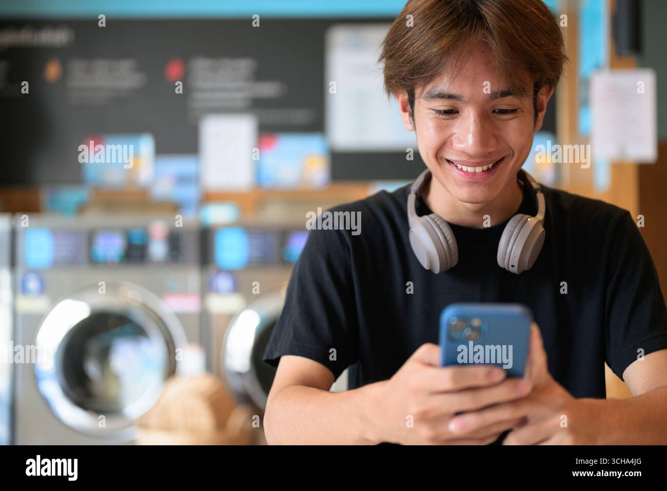 Giovane e affascinante uomo thailandese seduto in una moderna lavanderia con lavatrice che usa il telefono e indossa una t-shirt nera. Ritratto sincero che mostra la vita della Gen Z Foto Stock