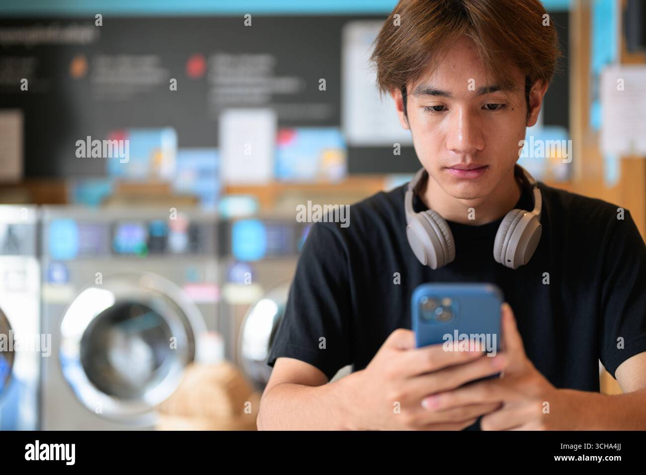 Giovane e affascinante uomo thailandese seduto in una moderna lavanderia con lavatrice che usa il telefono e indossa una t-shirt nera. Ritratto sincero che mostra la vita della Gen Z Foto Stock