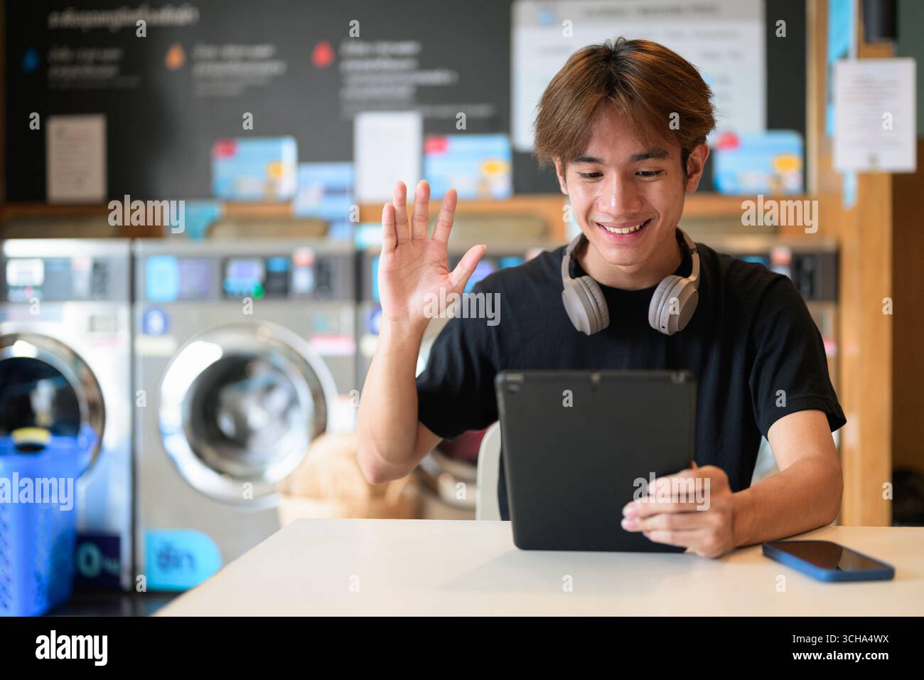 Giovane uomo thailandese seduto in una moderna lavanderia con lavatrice e con una t-shirt nera. Ritratto sincero che mostra lo stile di vita della generazione Z, le faccende quotidiane, Foto Stock