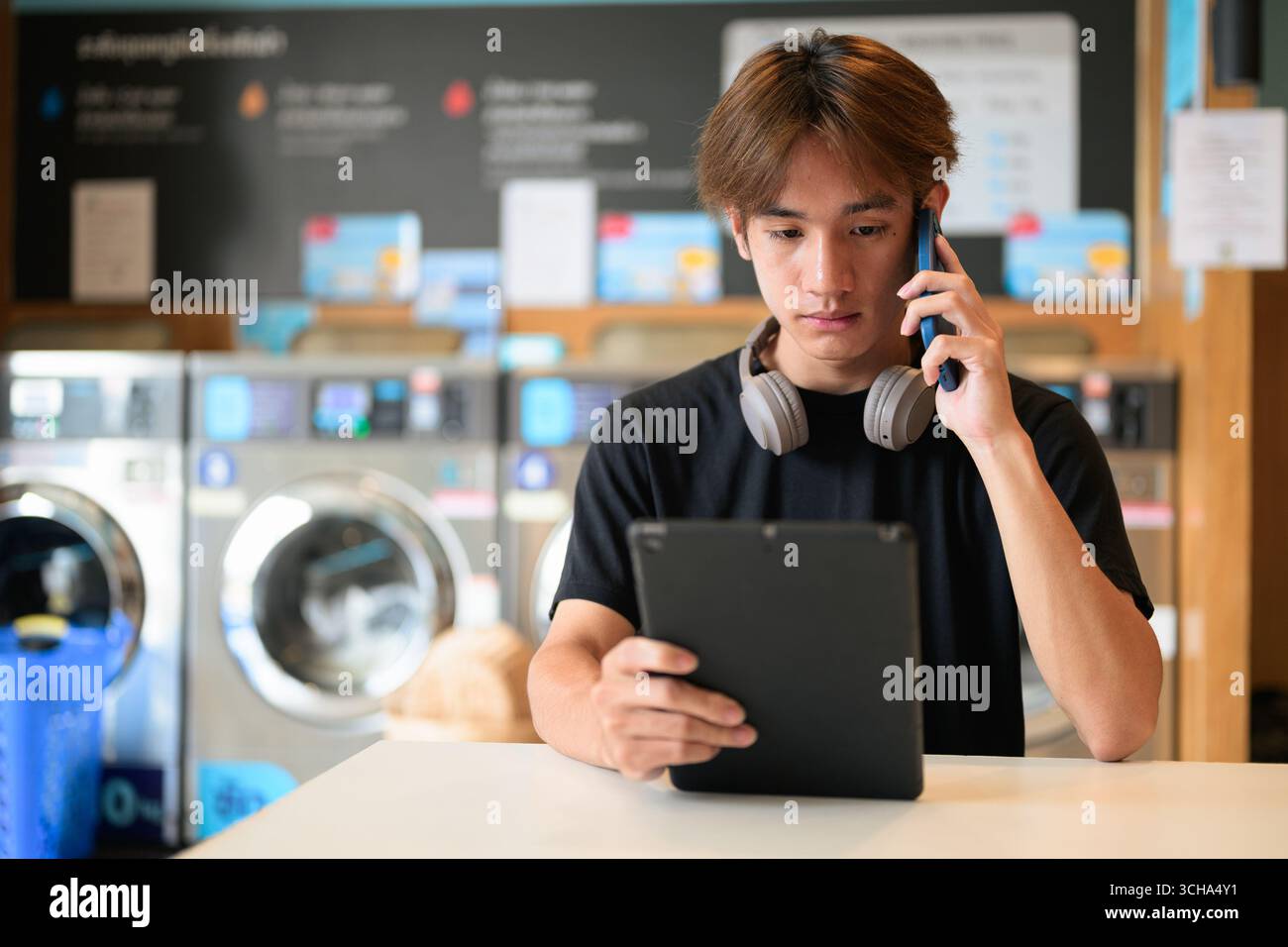 Giovane uomo thailandese seduto in una moderna lavanderia con lavatrice e con una t-shirt nera. Ritratto sincero che mostra lo stile di vita della generazione Z, le faccende quotidiane, Foto Stock