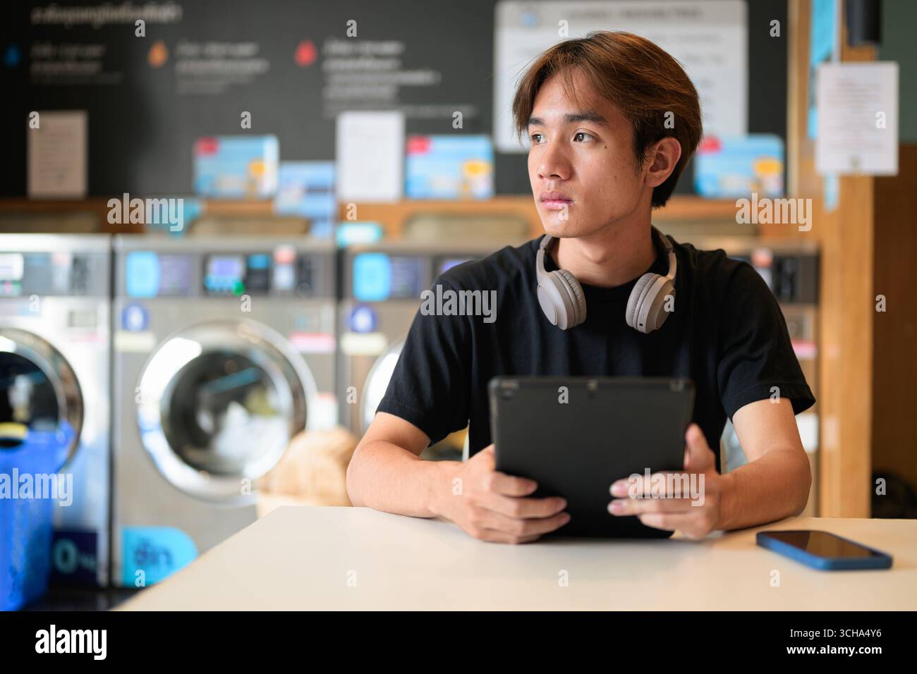 Giovane uomo thailandese seduto in una moderna lavanderia con lavatrice e con una t-shirt nera. Ritratto sincero che mostra lo stile di vita della generazione Z, le faccende quotidiane, Foto Stock