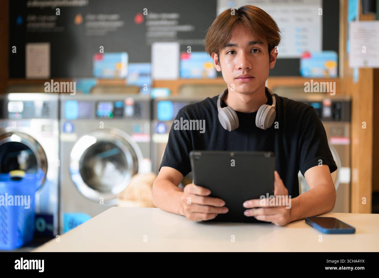 Giovane uomo thailandese seduto in una moderna lavanderia con lavatrice e con una t-shirt nera. Ritratto sincero che mostra lo stile di vita della generazione Z, le faccende quotidiane, Foto Stock