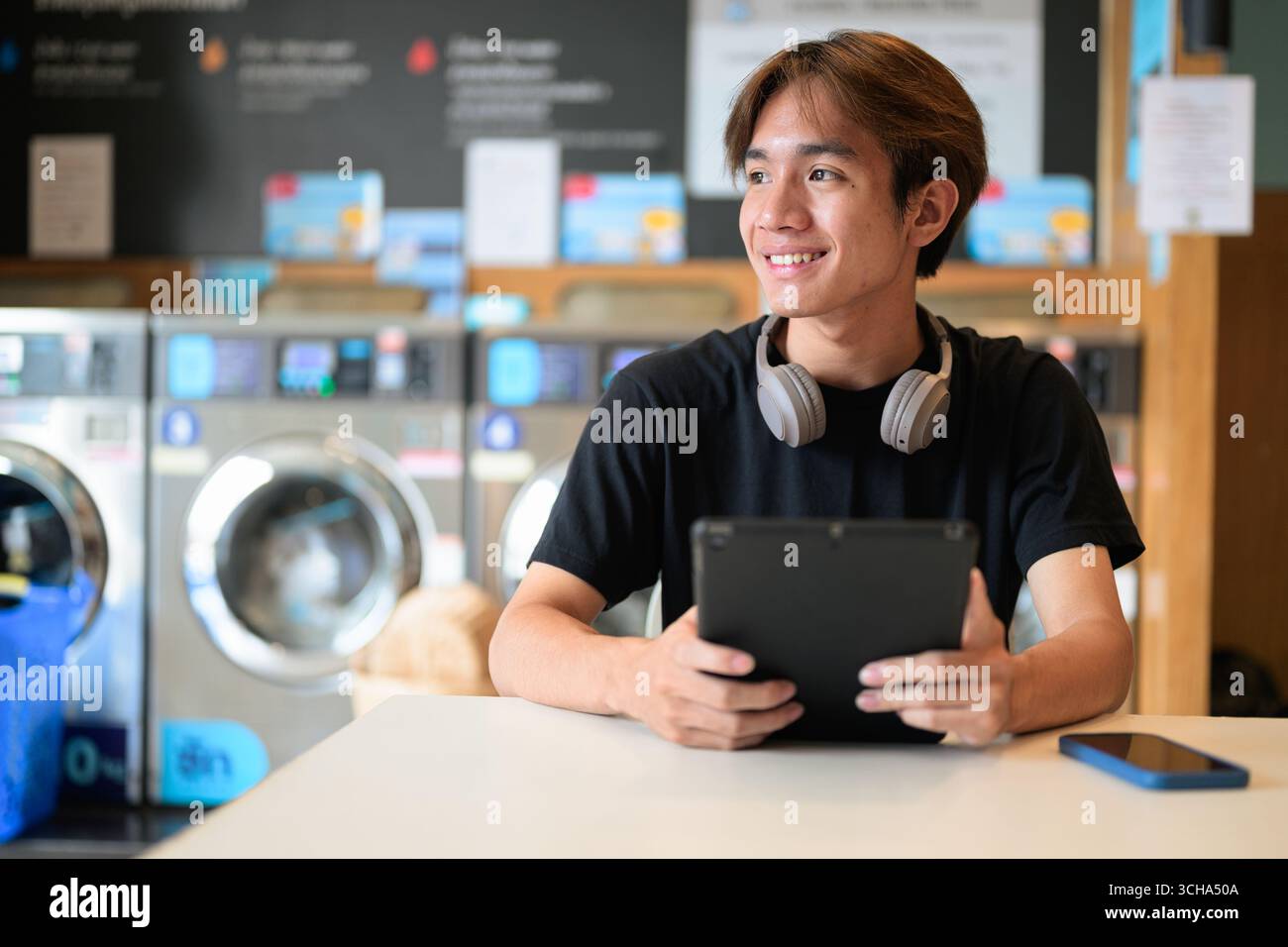 Giovane uomo thailandese seduto in una moderna lavanderia con lavatrice e con una t-shirt nera. Ritratto sincero che mostra lo stile di vita della generazione Z, le faccende quotidiane, Foto Stock