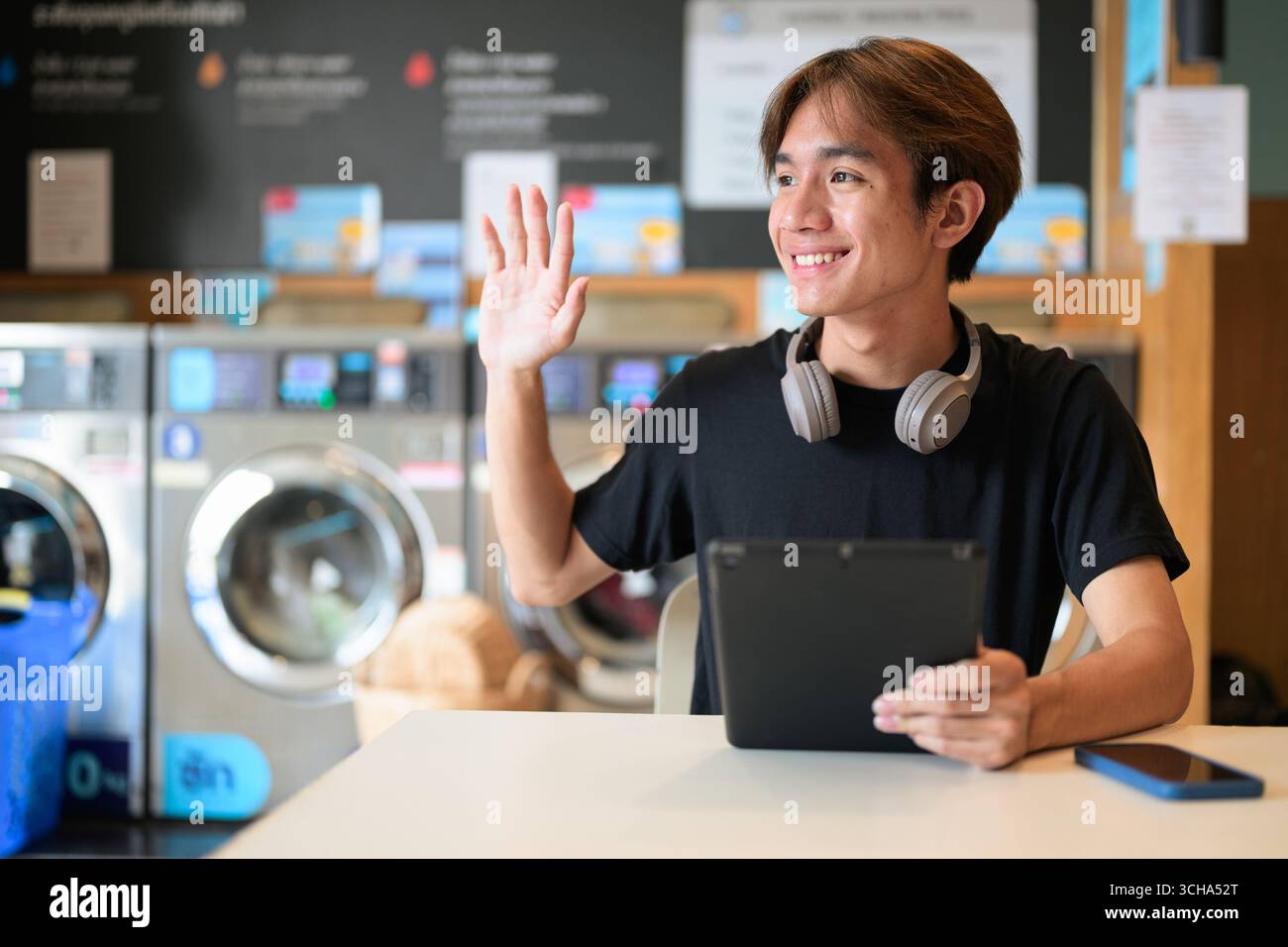 Giovane uomo thailandese seduto in una moderna lavanderia con lavatrice e con una t-shirt nera. Ritratto sincero che mostra lo stile di vita della generazione Z, le faccende quotidiane, Foto Stock