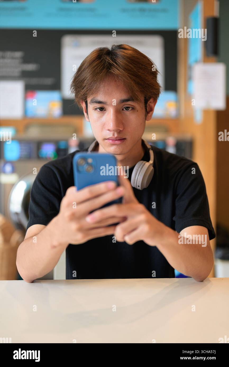 Giovane e affascinante uomo thailandese seduto in una moderna lavanderia con lavatrice che usa il telefono e indossa una t-shirt nera. Ritratto sincero che mostra la vita della Gen Z Foto Stock
