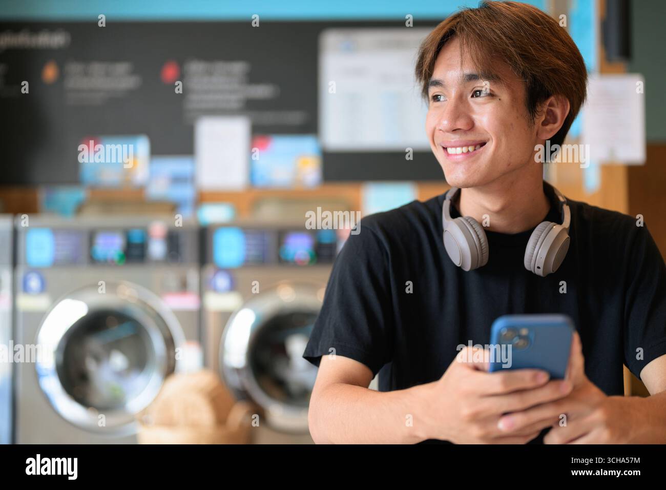 Giovane e affascinante uomo thailandese seduto in una moderna lavanderia con lavatrice che usa il telefono e indossa una t-shirt nera. Ritratto sincero che mostra la vita della Gen Z Foto Stock