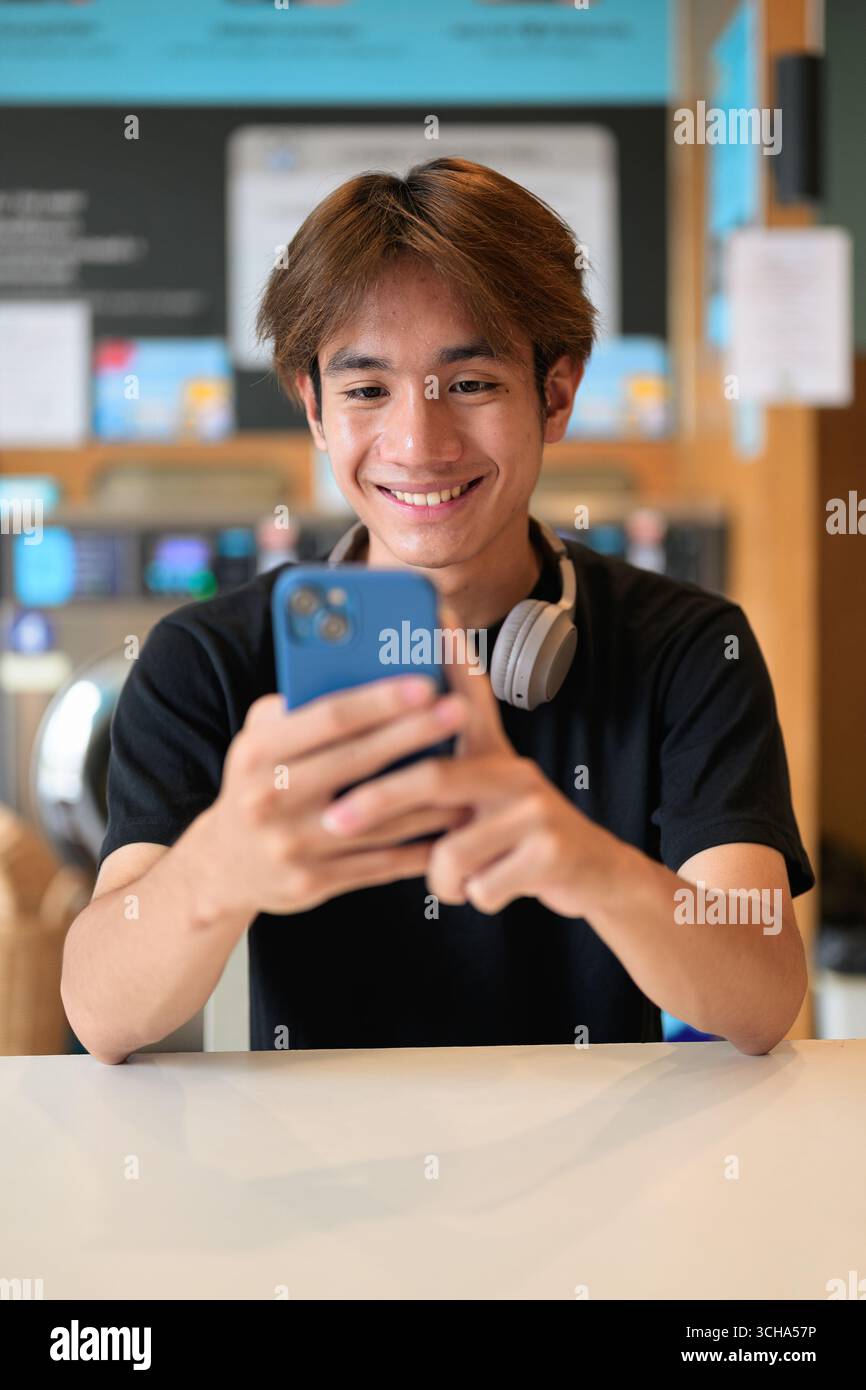 Giovane e affascinante uomo thailandese seduto in una moderna lavanderia con lavatrice che usa il telefono e indossa una t-shirt nera. Ritratto sincero che mostra la vita della Gen Z Foto Stock