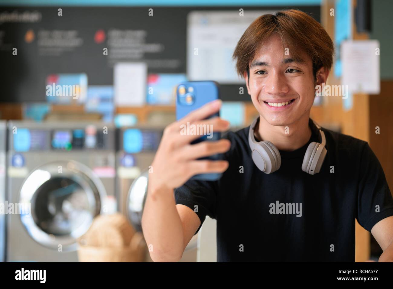 Giovane e affascinante uomo thailandese seduto in una moderna lavanderia con lavatrice che usa il telefono e indossa una t-shirt nera. Ritratto sincero che mostra la vita della Gen Z Foto Stock