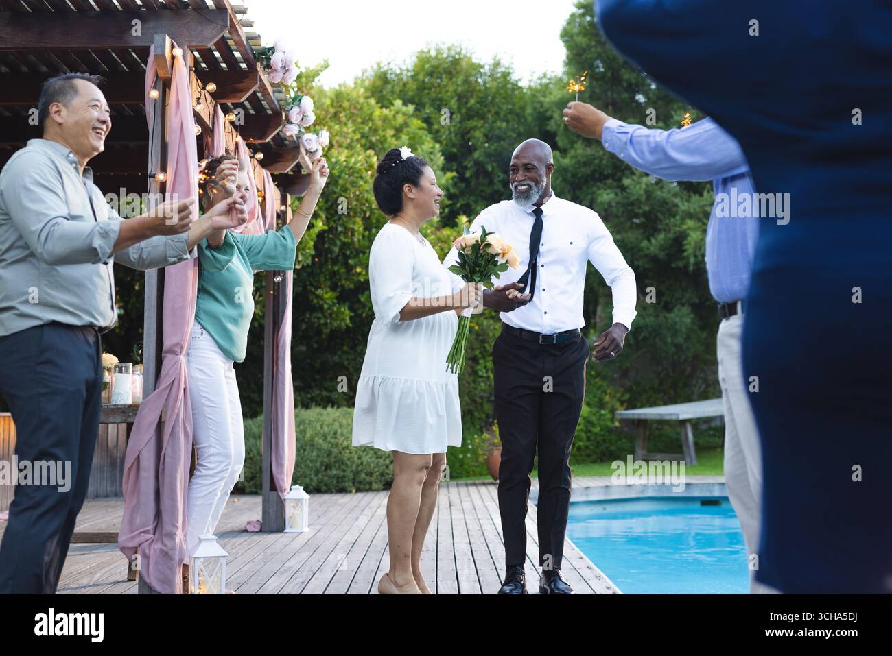 Al matrimonio, festeggiando a bordo piscina, coppia senior con amici che tengono scintillanti, sorridendo gioiosamente. Festa, festa, festa, amicizia, felicità Foto Stock
